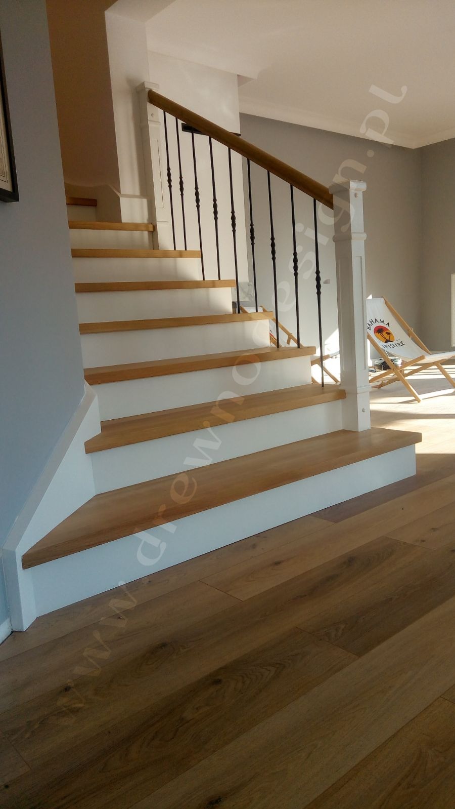 Drewniane schody z białymi podstopnicami i czarną, kutą balustradą, widoczne z dołu, z jasną podłogą i leżakiem 'Bahama House' w tle.