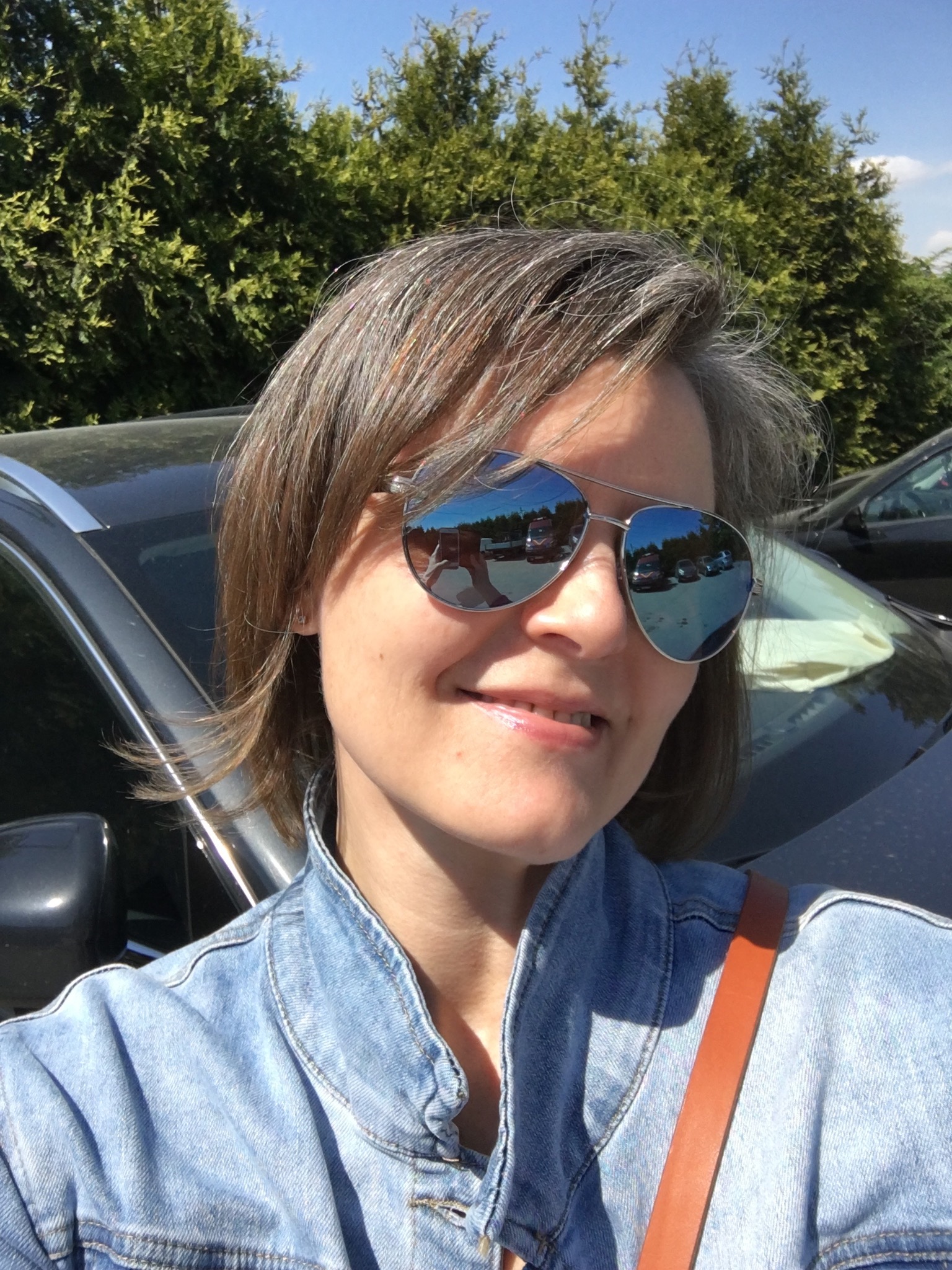 Portret kobiety w okularach przeciwsłonecznych, odbijających parking z samochodami, stojącej przed czarnym SUV-em w słoneczny dzień.