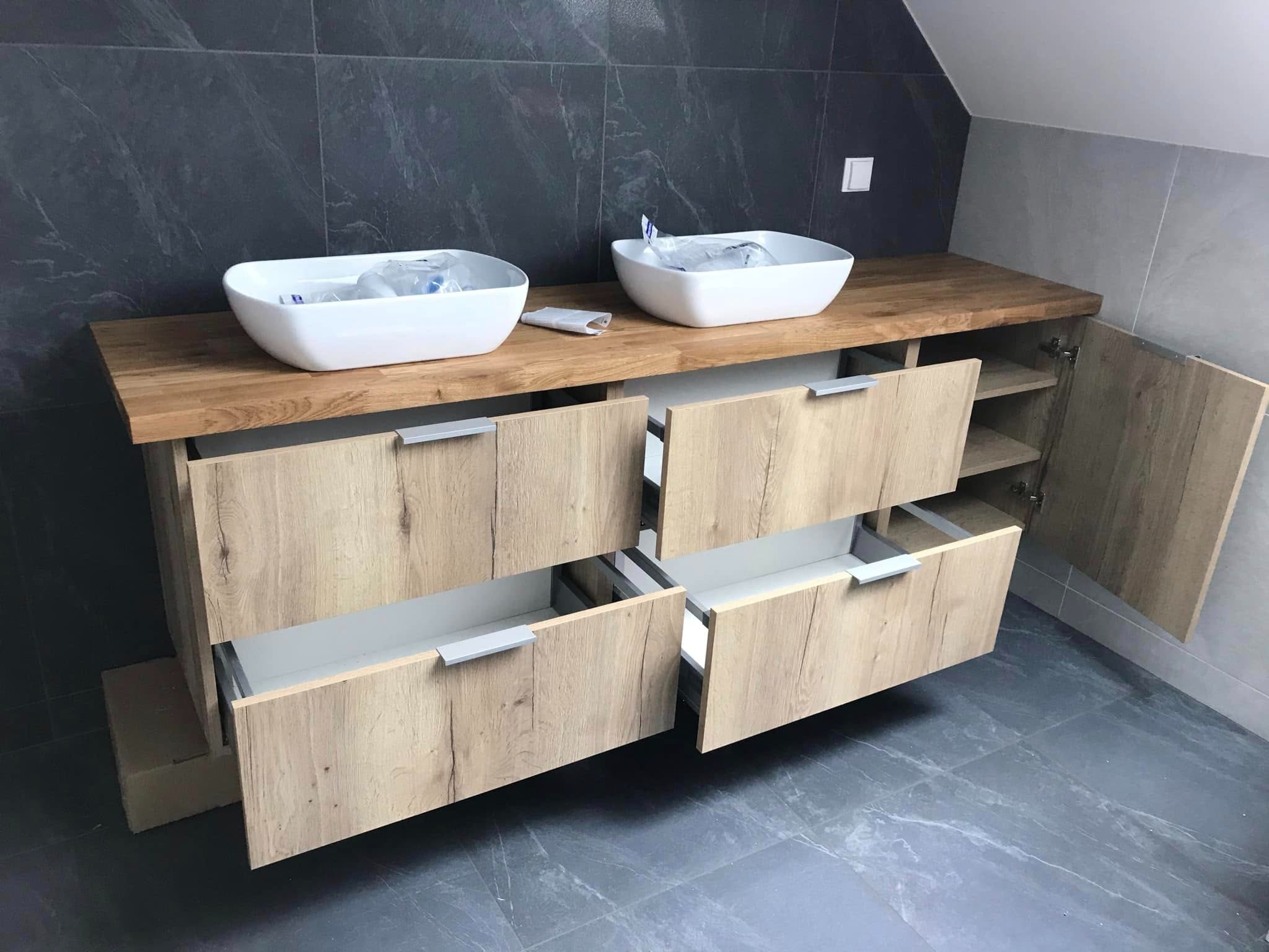 Nowoczesna szafka łazienkowa z blatem z jasnego drewna, dwie białe umywalki nablatowe, otwarte szuflady i szafka z półkami. Szare płytki na ścianie i podłodze.