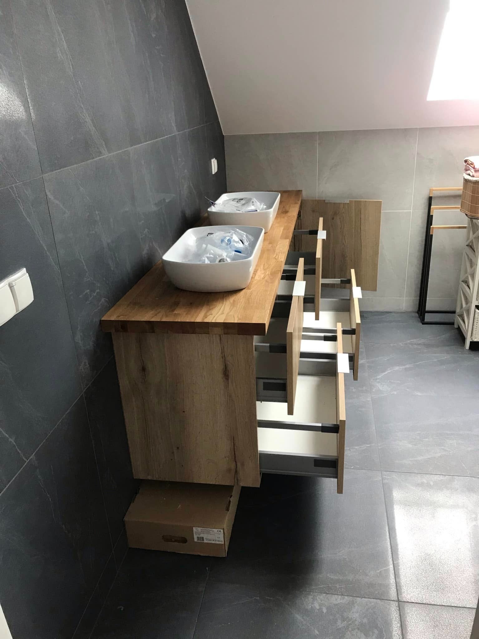 Drewniana szafka łazienkowa z dwiema umywalkami i wysuniętymi szufladami, na tle szarych płytek. Widoczny karton pod szafką i fragment wyposażenia łazienki.