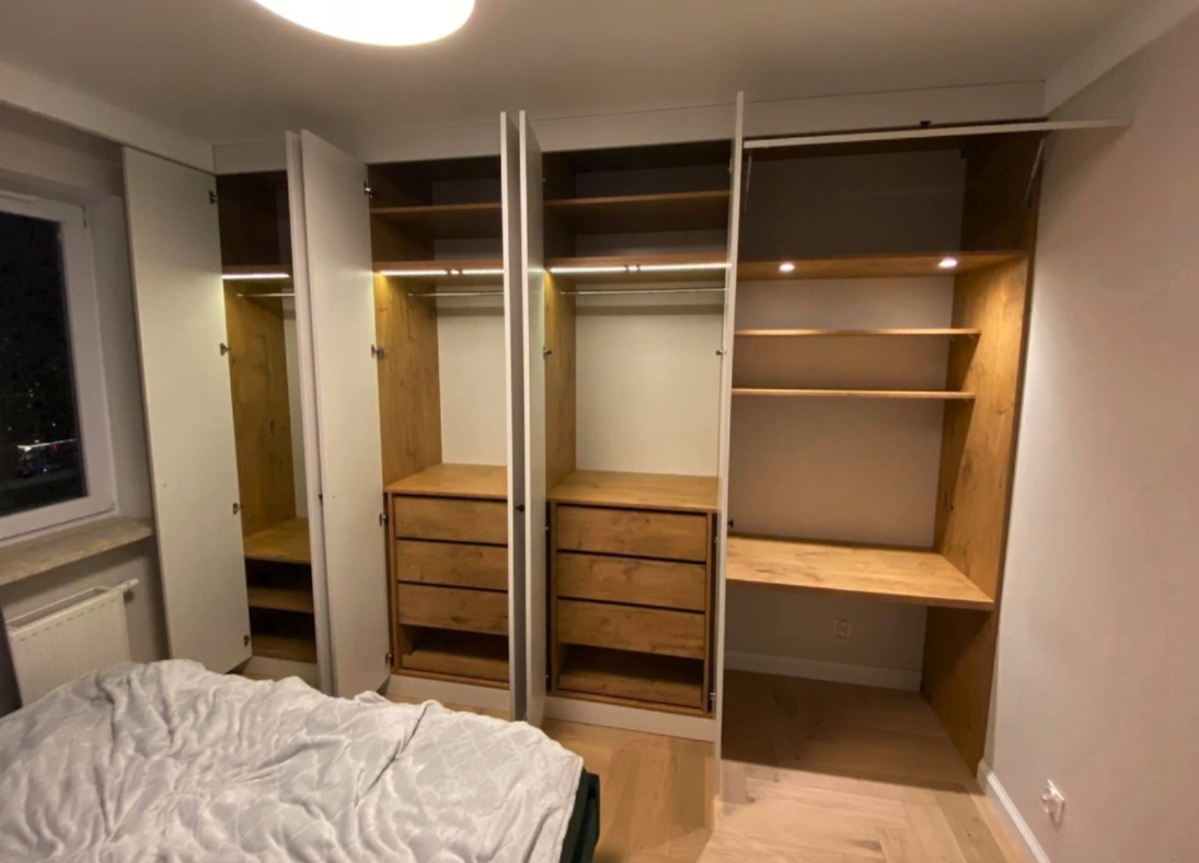 Nowoczesna szafa wnękowa z drewnianymi elementami i oświetleniem LED, widoczna z perspektywy wnętrza sypialni, z częściowo otwartymi drzwiami.