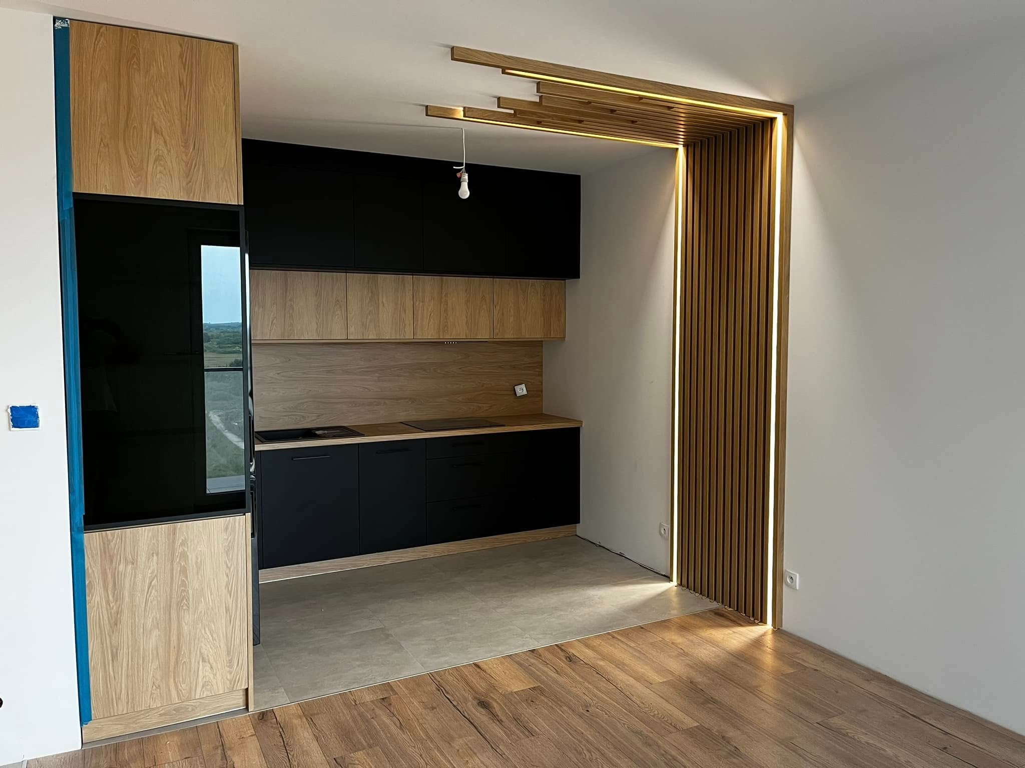 Nowoczesna kuchnia z czarnymi szafkami i drewnianymi frontami, oświetleniem LED w dekoracyjnej obudowie przejścia. Widok na drewnianą podłogę i fragment okna.
