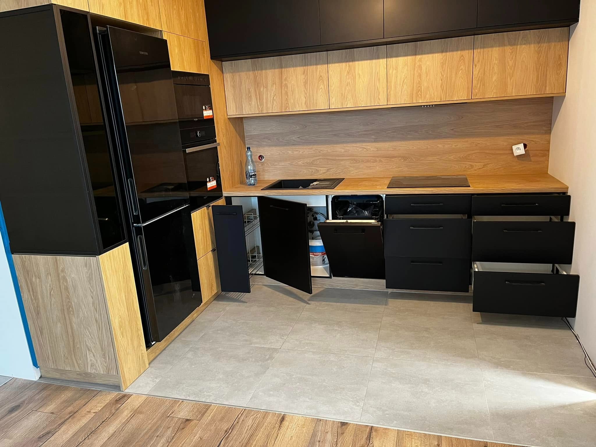 Nowoczesna kuchnia z czarnymi frontami i drewnianymi akcentami. Otwarta szafka ukazuje metalowe półki. Widoczny sprzęt AGD w zabudowie i szare płytki podłogowe.