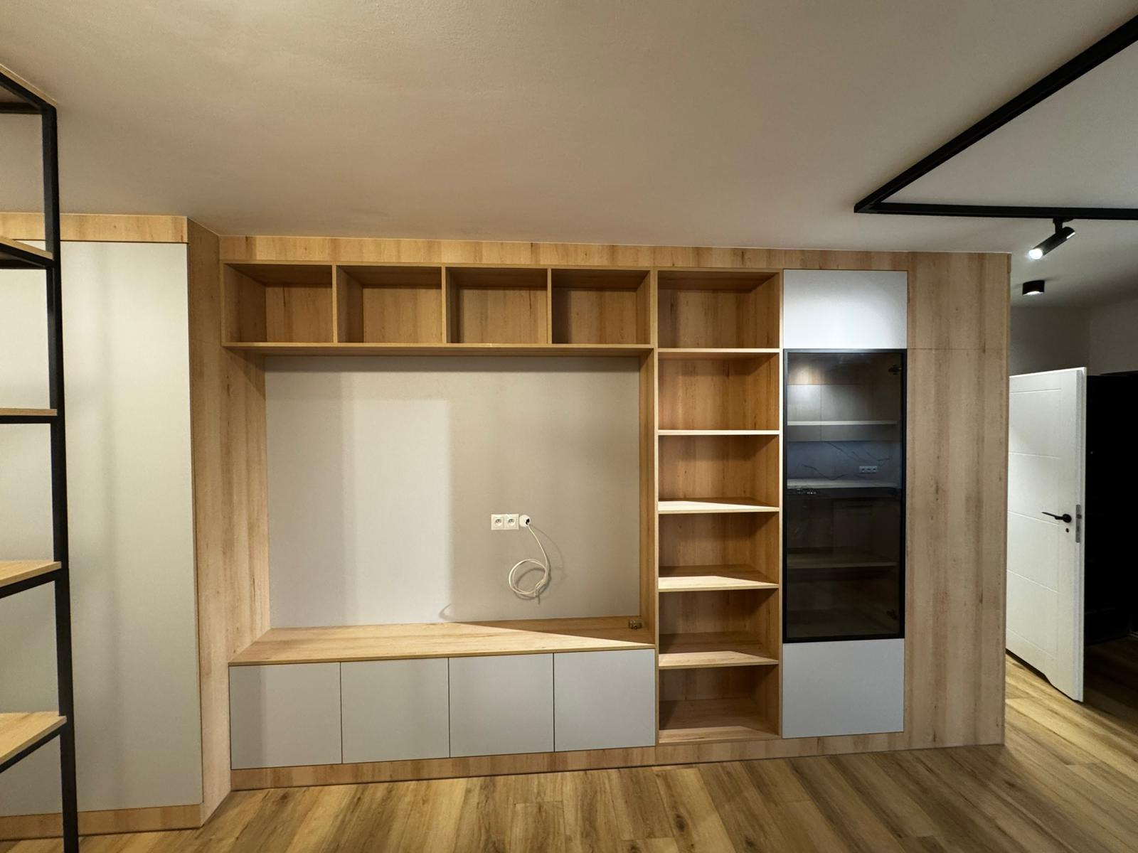 Drewniana zabudowa ściany z półkami, szafkami i przeszkloną witryną w nowoczesnym salonie. Widoczne gniazdka elektryczne i fragment podłogi.