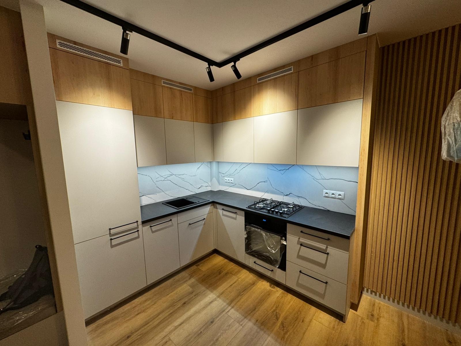 Nowoczesna kuchnia narożna z białymi i drewnianymi frontami, czarnym blatem i marmurowym backsplashiem. Oświetlenie podszafkowe i punktowe. Parkiet na podłodze.