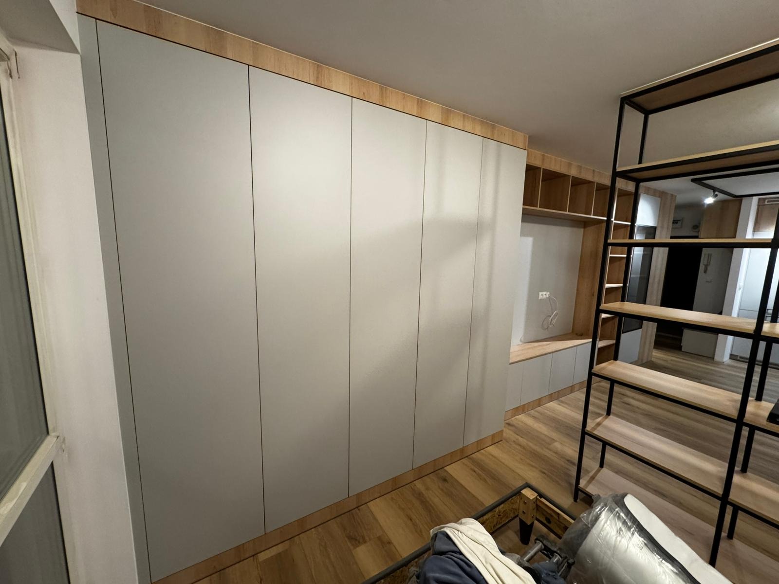 Jasnoszara zabudowa meblowa z drewnianymi akcentami, obejmująca szafy i półki, uzupełniona metalowym regałem w nowoczesnym wnętrzu. Widoczna podłoga z paneli.