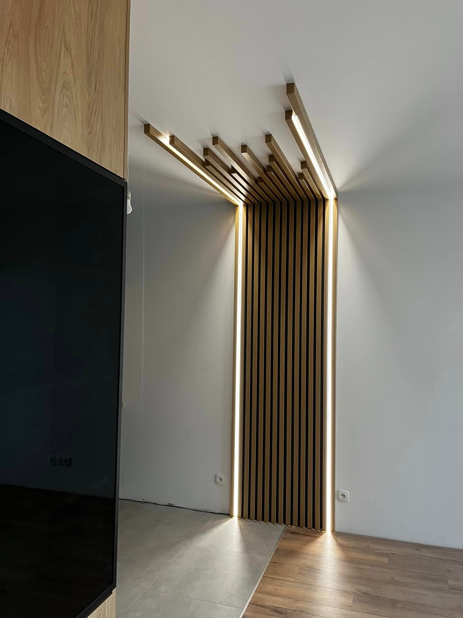 Drewniane lamele ścienne z podświetleniem LED tworzą nowoczesny akcent w jasnym wnętrzu. Widoczne fragmenty podłogi i telewizora.