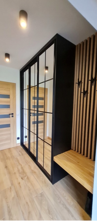 Czarna szafa wnękowa z lustrzanymi drzwiami przesuwnymi i drewnianym siedziskiem w przedpokoju. Widoczne drzwi wejściowe i oświetlenie sufitowe.