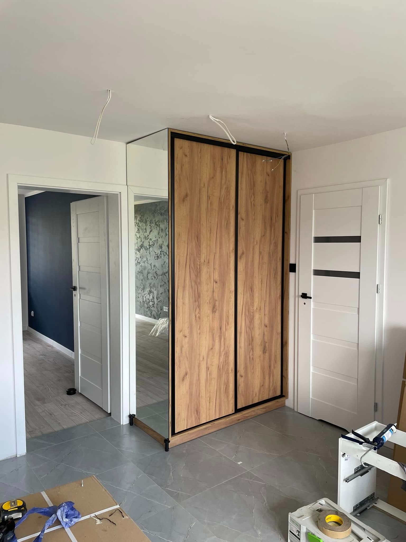 Nowoczesna szafa przesuwna z lustrem i drewnianymi frontami, czarne obramowanie, szare płytki podłogowe, widok na wnętrze pokoju z otwartymi drzwiami.