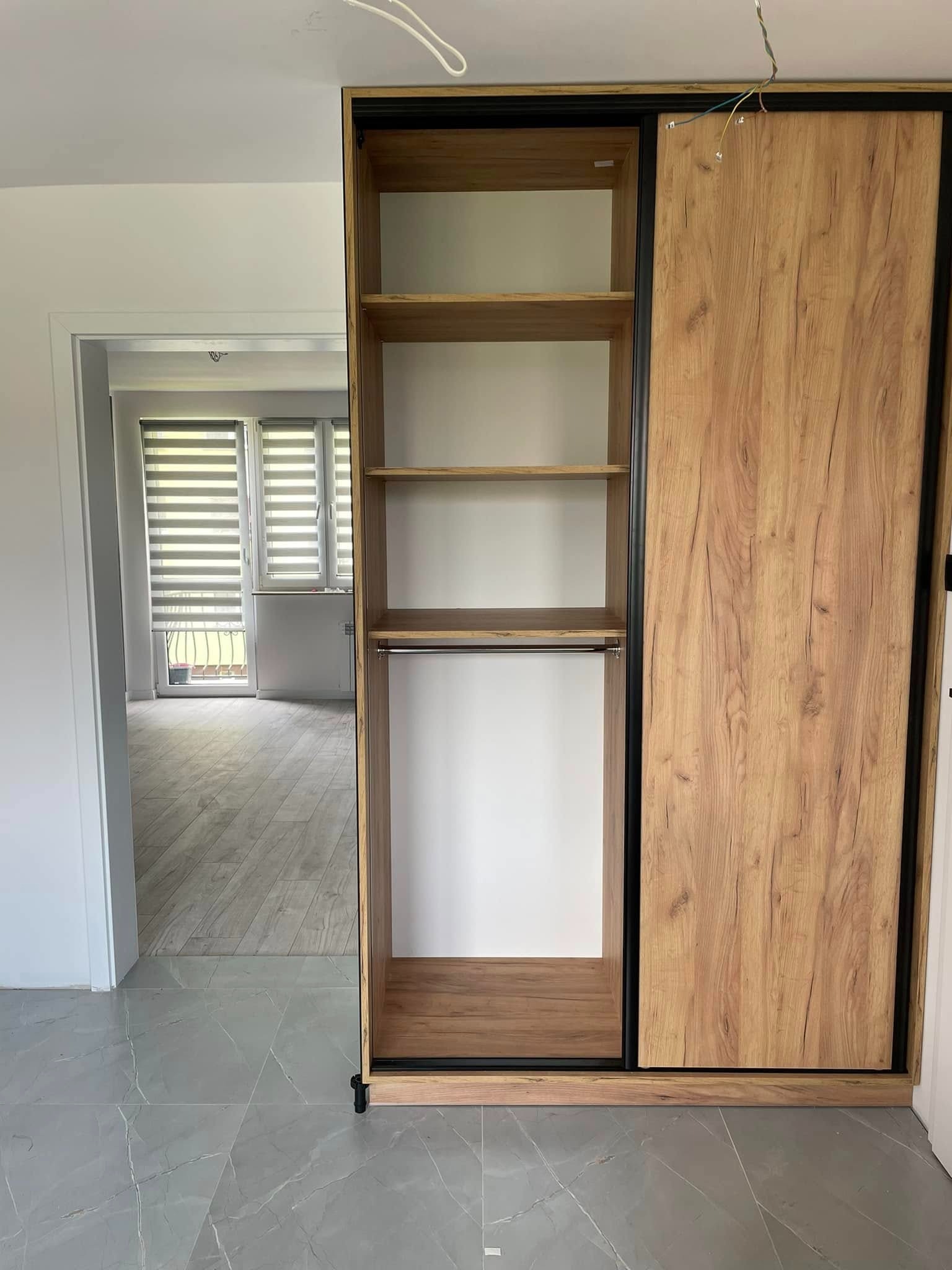 Szafa przesuwna z drewnopodobnymi frontami i czarnymi obramowaniami, widok na półki i drążek, w tle okno z żaluzjami, szare płytki podłogowe.