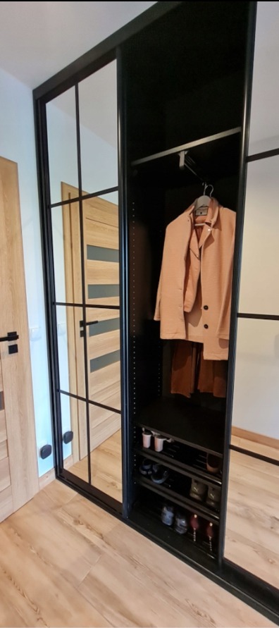 Czarna szafa wnękowa z przesuwnymi drzwiami z lustrami i czarnymi szprosami. Wnętrze szafy z ubraniami i półkami na buty. Widoczna drewniana podłoga i drzwi.
