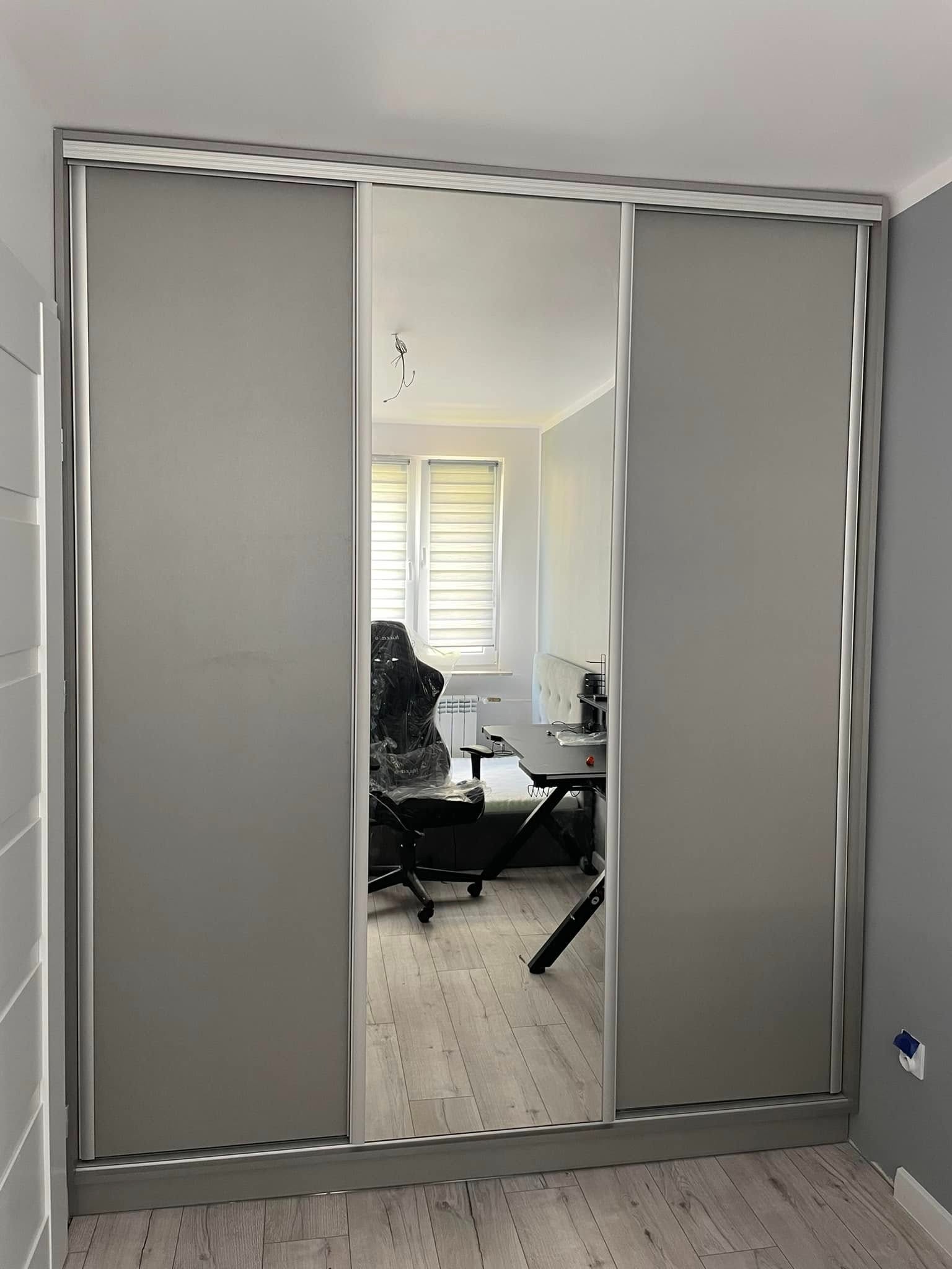 Szara szafa przesuwna z lustrem, odbijająca wnętrze pokoju z biurkiem i krzesłem. Minimalistyczny design, jasne wnętrze, widoczne detale wykończenia.
