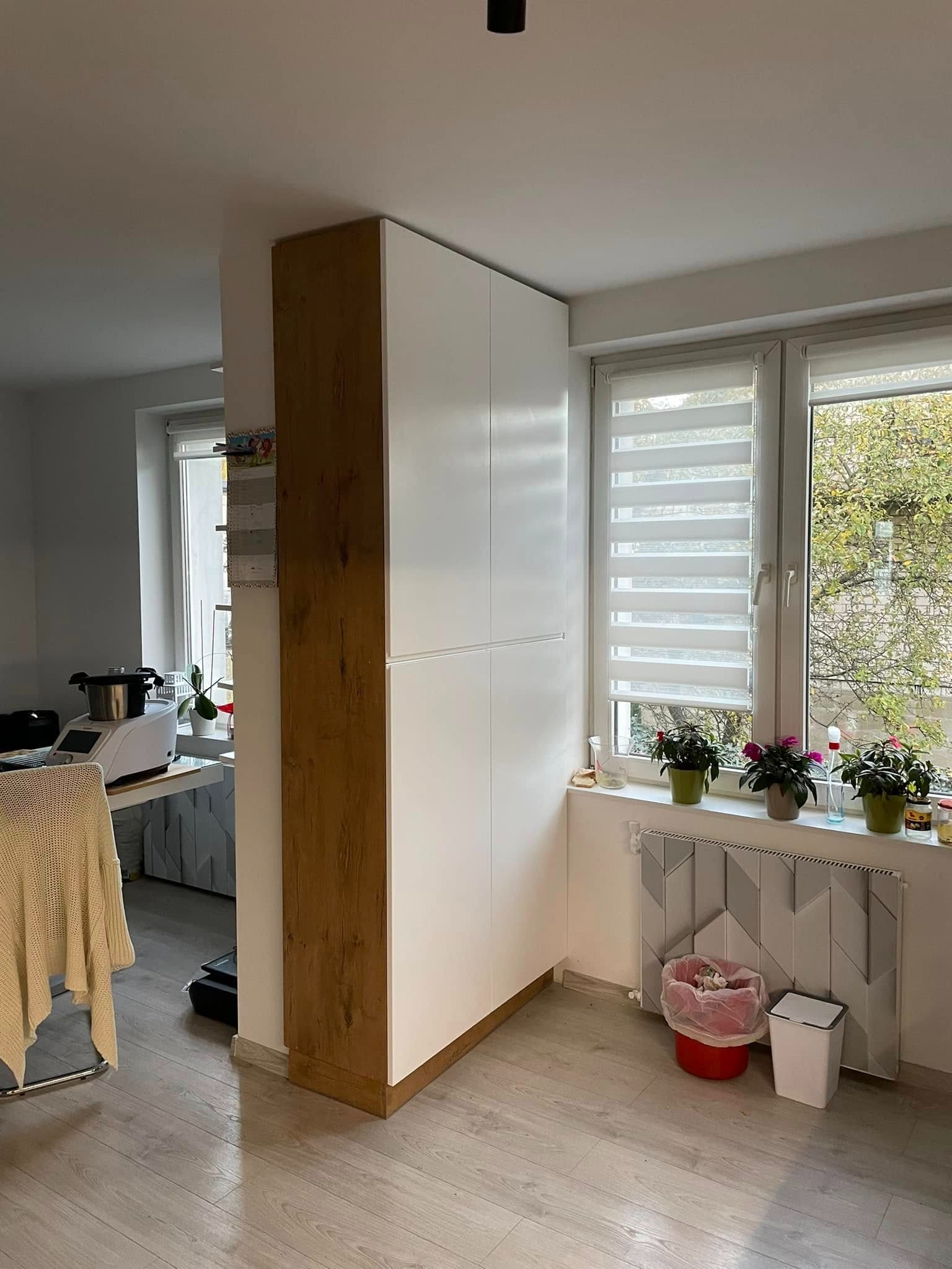 Wysoka, zabudowana szafa z białymi frontami i drewnianym bokiem, wkomponowana w przestrzeń między ścianą a oknem. Widoczne kwiaty na parapecie i grzejnik z geometrycznym wzorem.