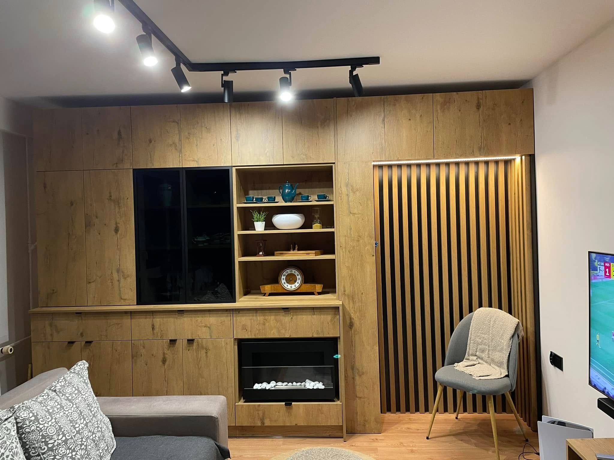 Drewniana zabudowa ściany w salonie z czarnymi akcentami: przeszklona szafka, półki z dekoracjami, imitacja kominka i pionowe lamele. Oświetlenie punktowe.