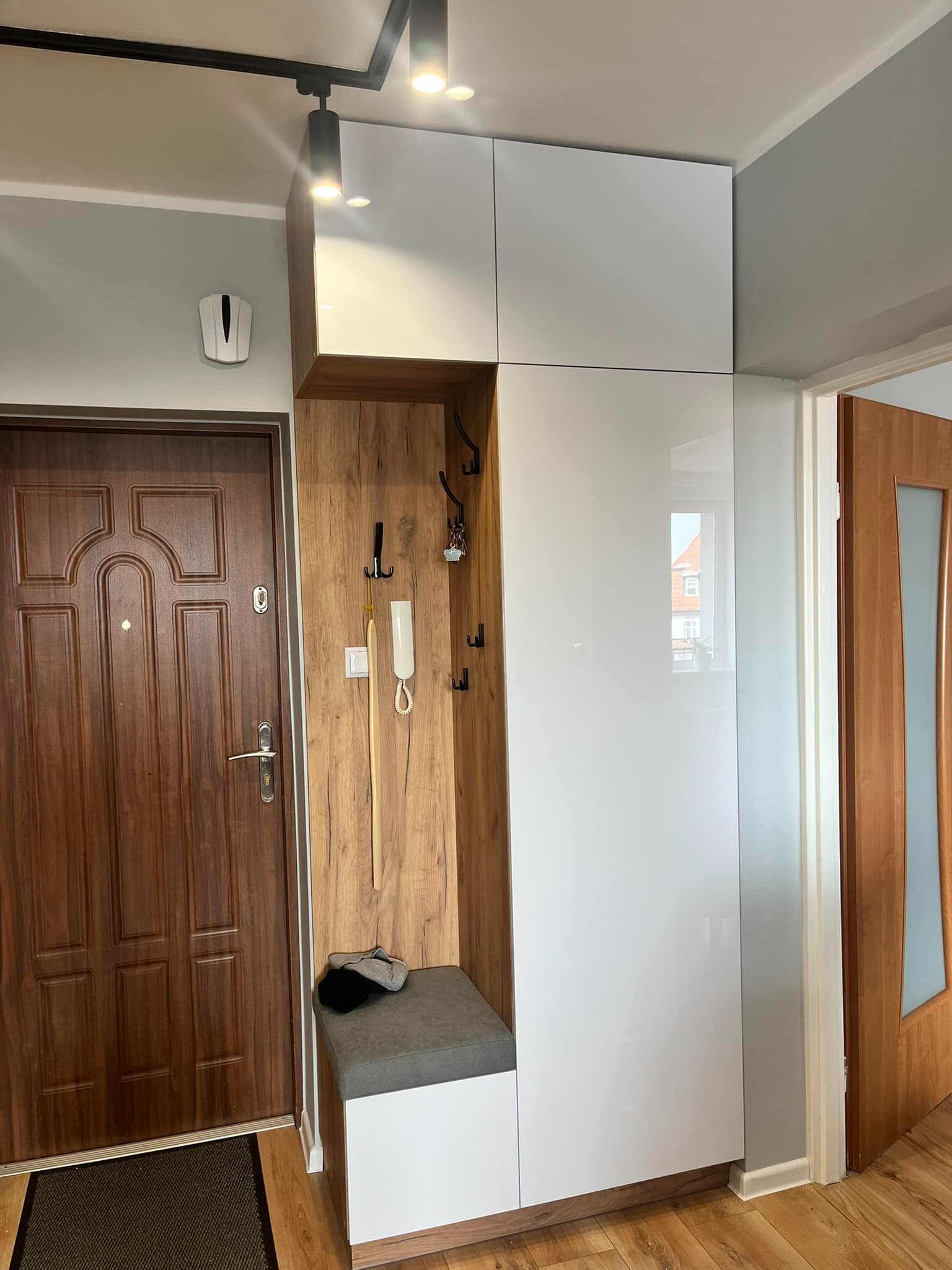 Funkcjonalna zabudowa przedpokoju z siedziskiem, wieszakami i szafkami z białymi, lakierowanymi frontami. Drewnopodobne wykończenie wnęki i dolnej części szafy.