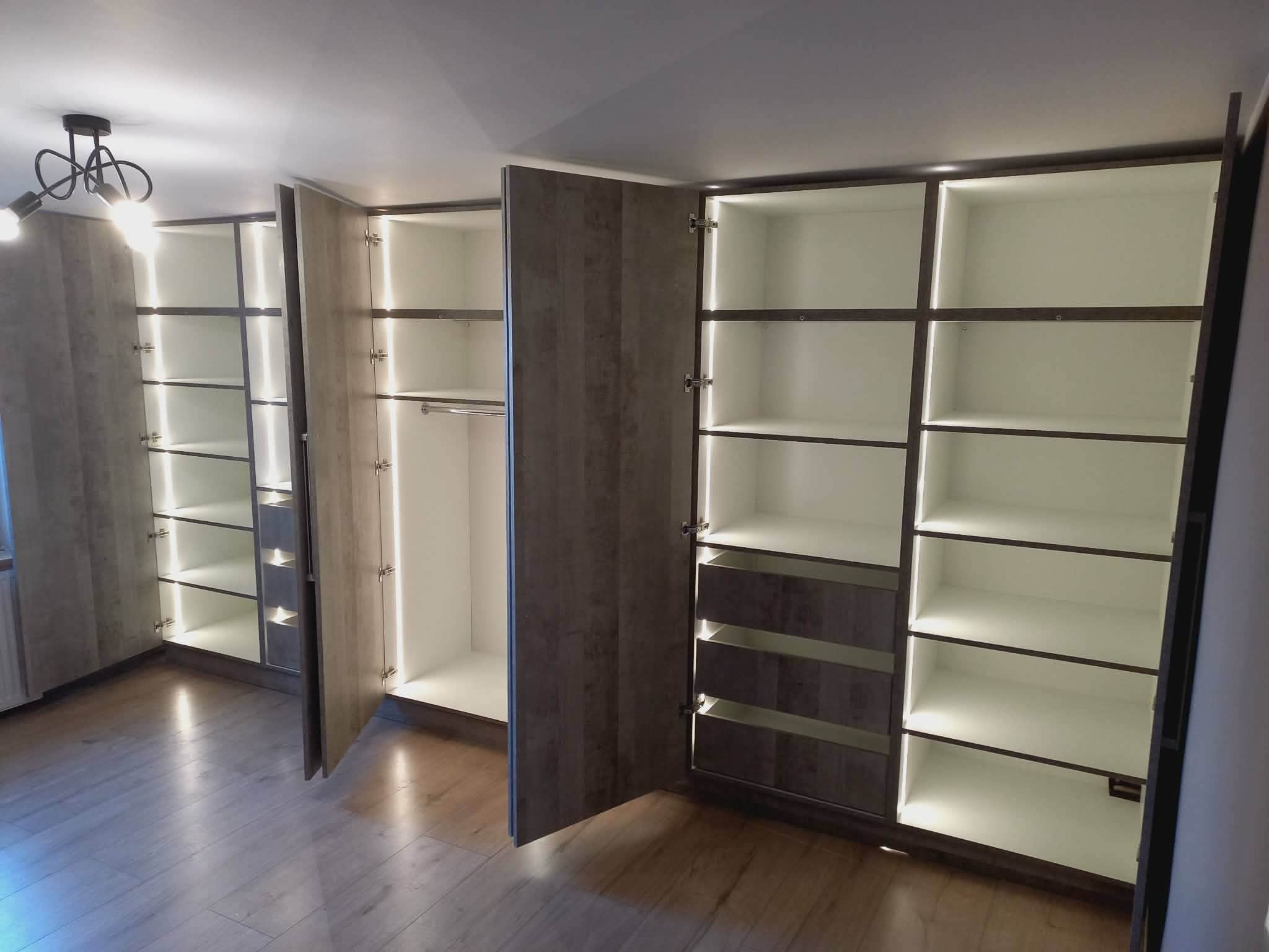 Nowoczesna szafa wnękowa z oświetleniem LED, otwarte drzwi ukazują półki i szuflady. Jasne wnętrze kontrastuje z ciemniejszymi obramowaniami i drewnopodobnymi frontami.