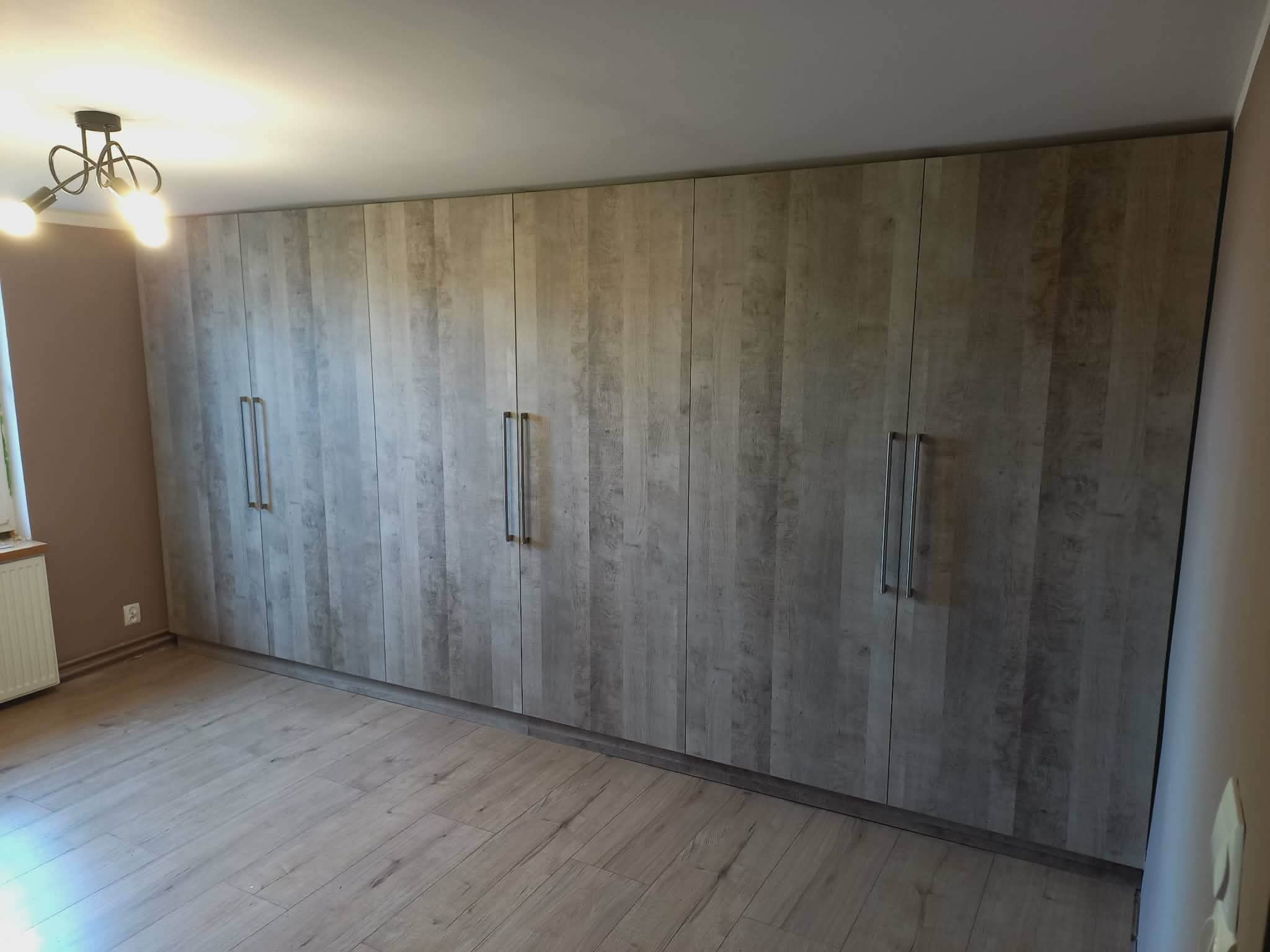 Duża, zabudowana szafa z frontami imitującymi drewno, z długimi, pionowymi uchwytami, zajmująca całą ścianę w pokoju z panelami podłogowymi w odcieniach szarości.