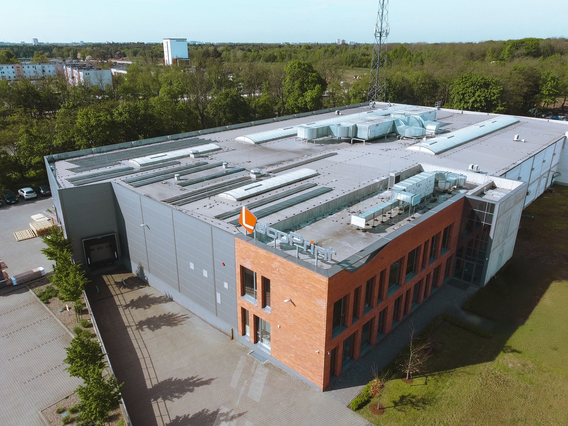 Widok z lotu ptaka na nowoczesny budynek przemysłowy z ceglaną elewacją i szarymi panelami, logo na dachu, liczne okna, systemy wentylacyjne na dachu, parking z paletami i zielenią w tle.