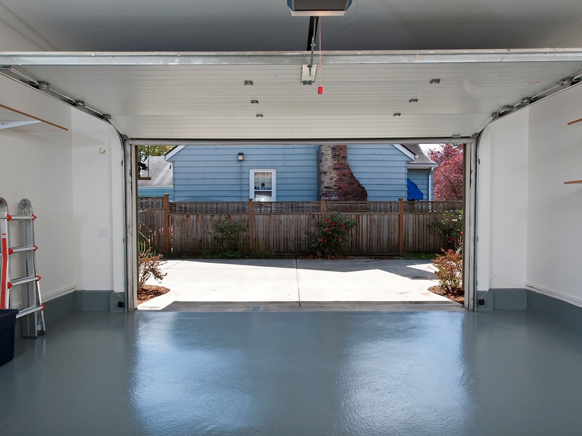 Wnętrze garażu z otwartą bramą, widok na podwórko z drewnianym ogrodzeniem i domem w tle, podłoga garażu pokryta szarą, błyszczącą farbą epoksydową, drabina oparta o ścianę po lewej stronie.