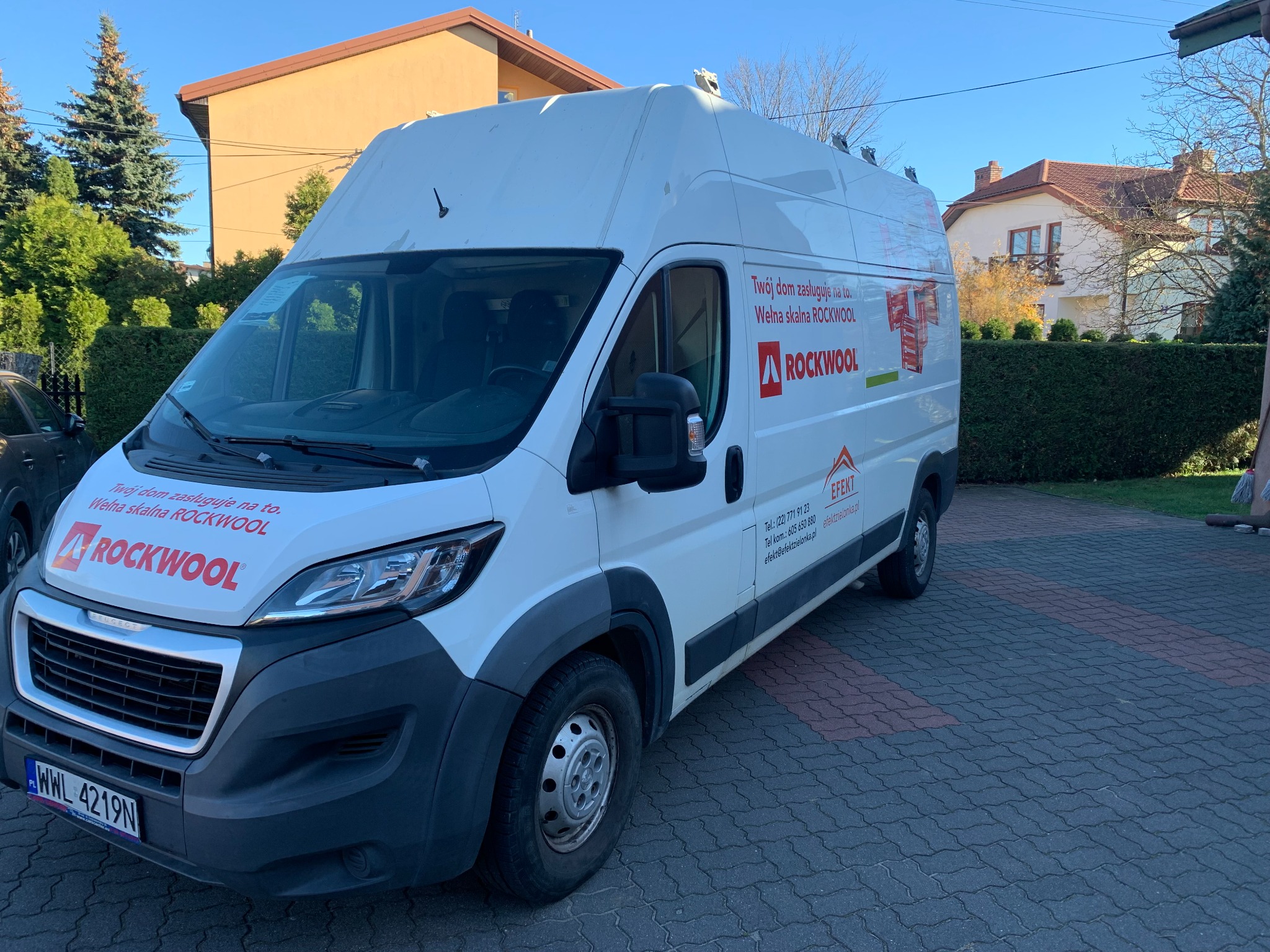 Biały samochód dostawczy z reklamą wełny mineralnej Rockwool zaparkowany na brukowanym podjeździe przed domem.