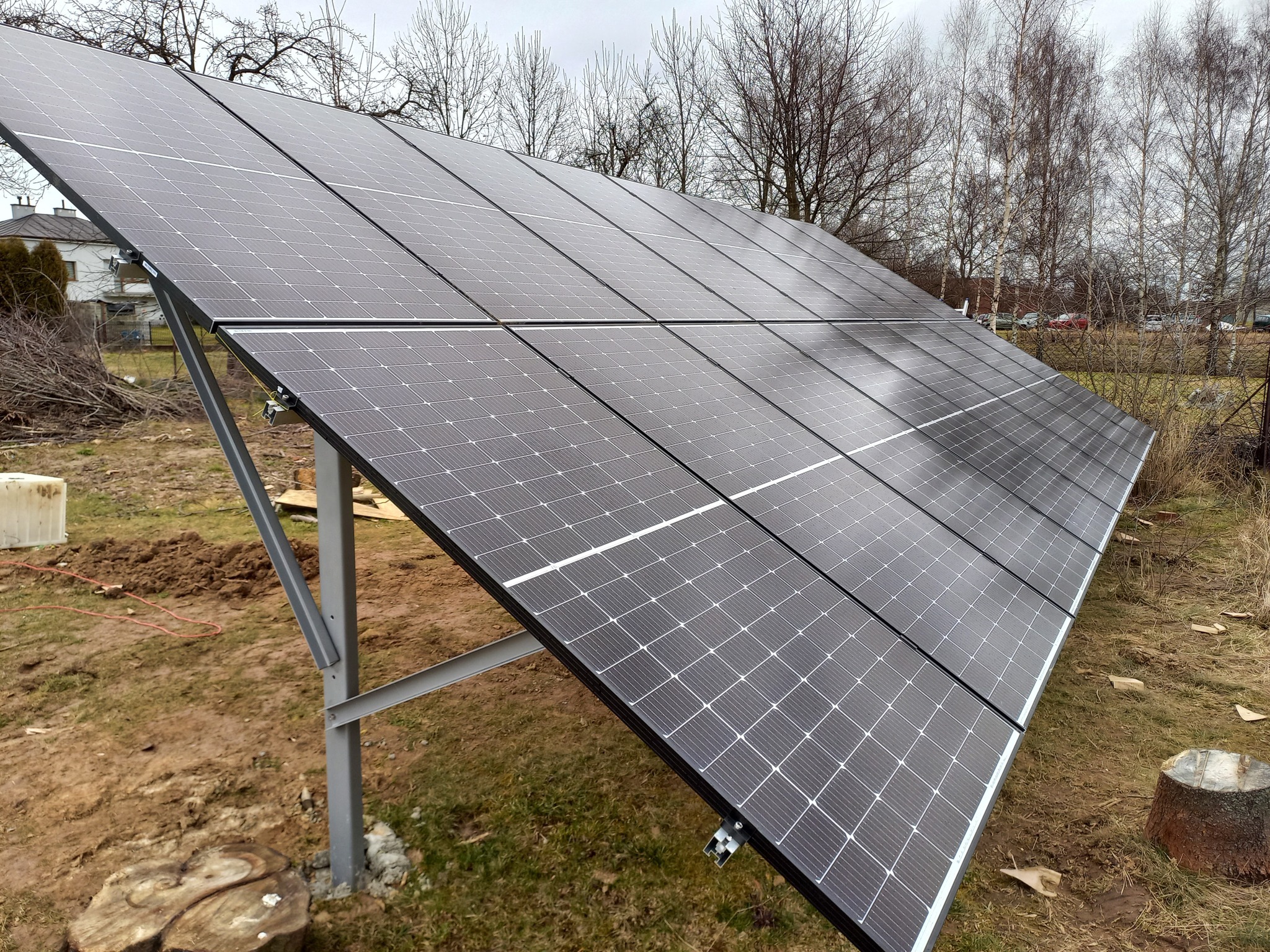 Rząd paneli słonecznych na metalowej konstrukcji, widoczne pnie drzew i budynek w tle.