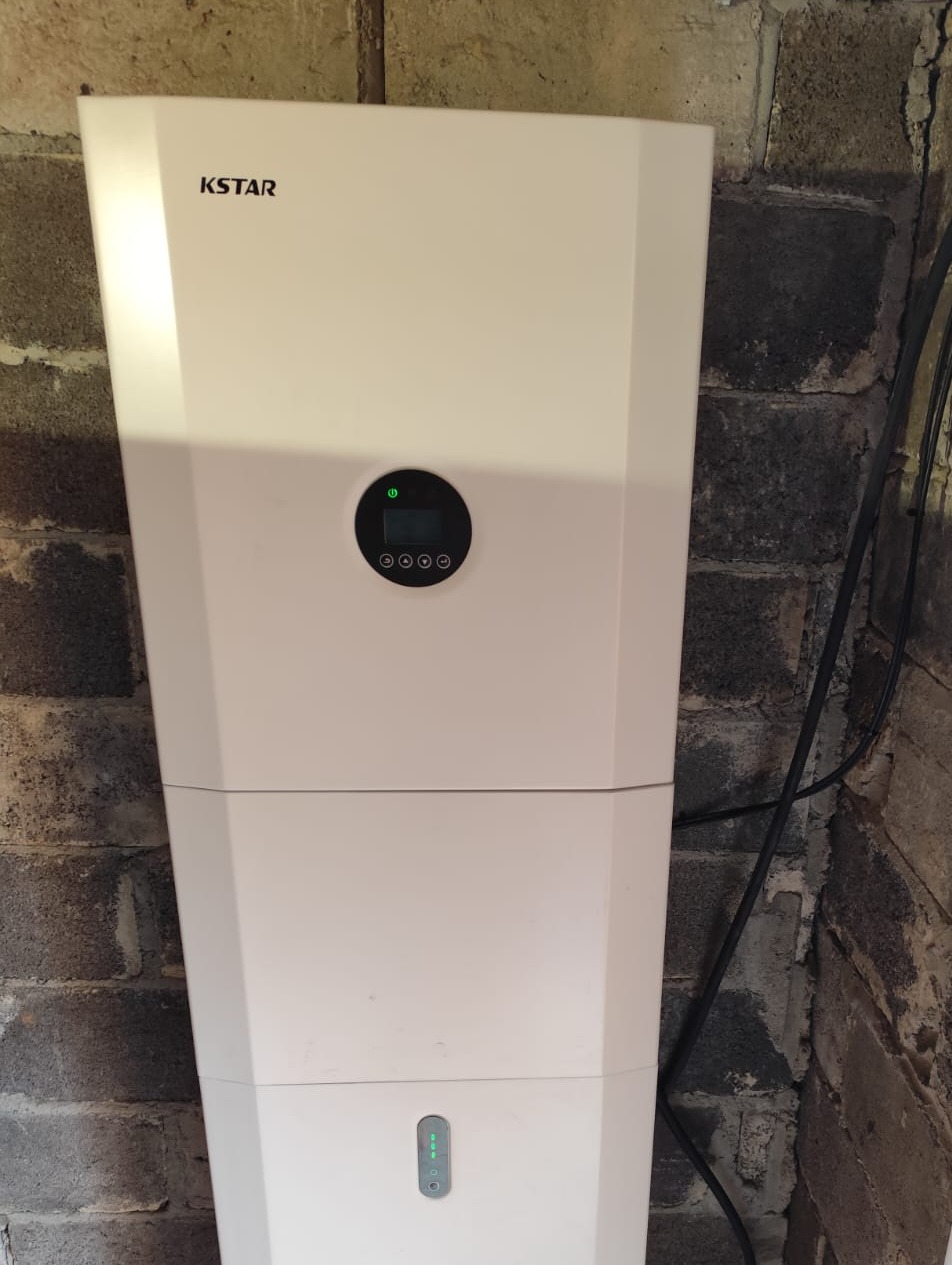 Biały moduł magazynu energii KSTAR zamontowany na tle surowej, ceglanej ściany z widocznymi przewodami.