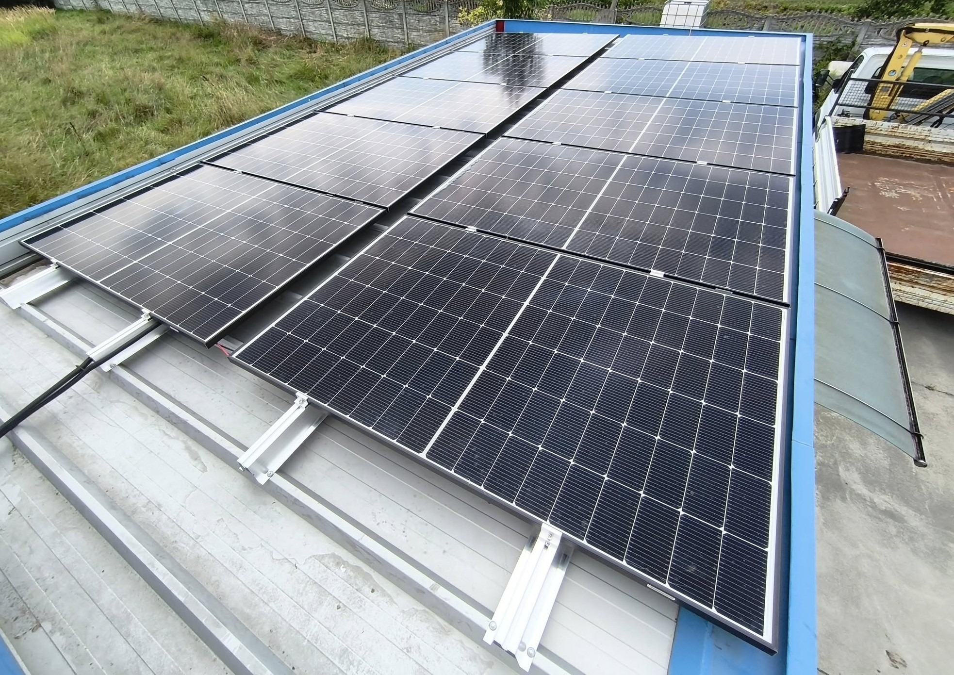 Panel słoneczny na dachu z blachy falistej, widok z góry. Montaż na aluminiowych profilach. Częściowo widoczny pojazd w tle. Dachy budynków i zieleń w tle.