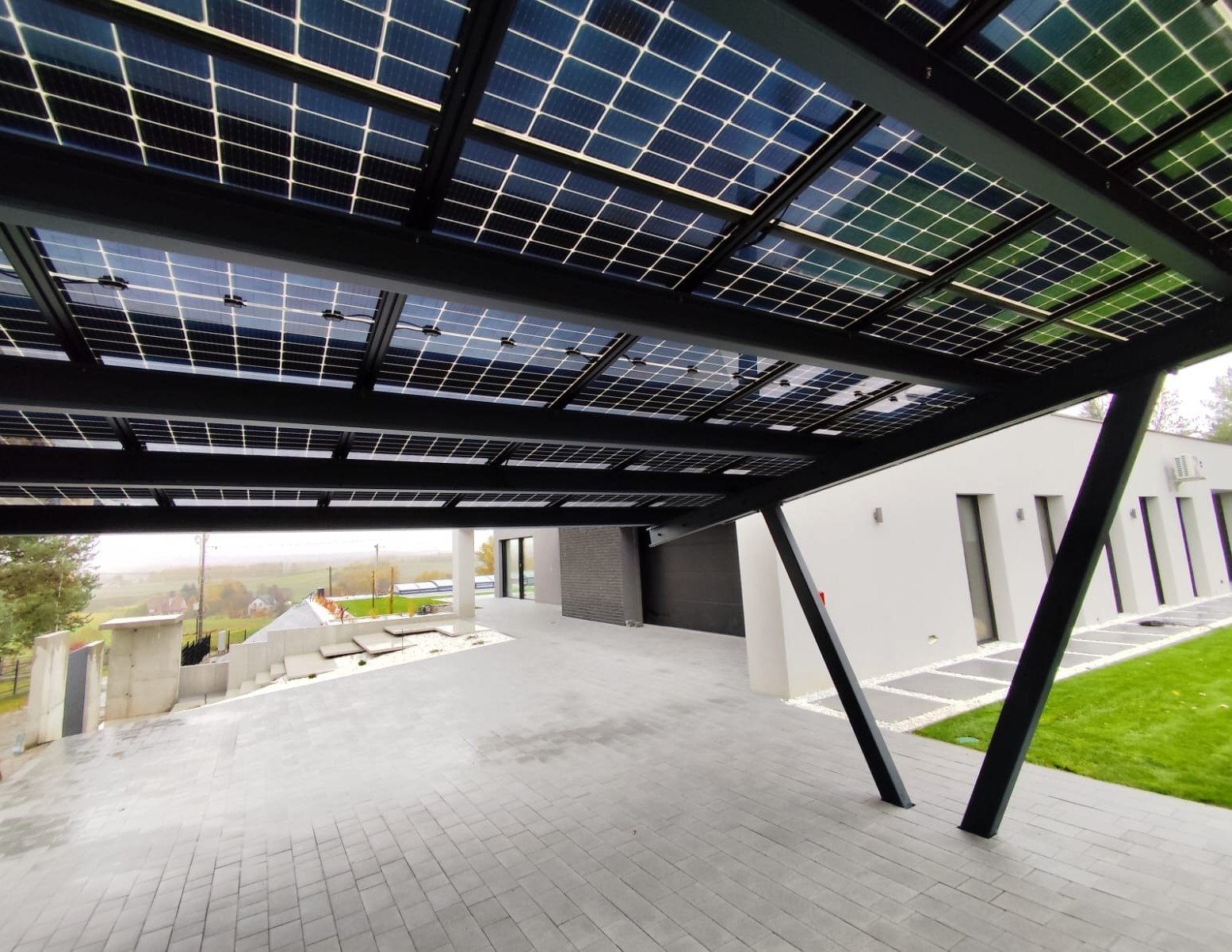 Zadaszenie z paneli słonecznych nad podjazdem, nowoczesny design. Widok z dołu, szara kostka brukowa, biały budynek w tle. Nietypowe rozwiązanie architektoniczne.