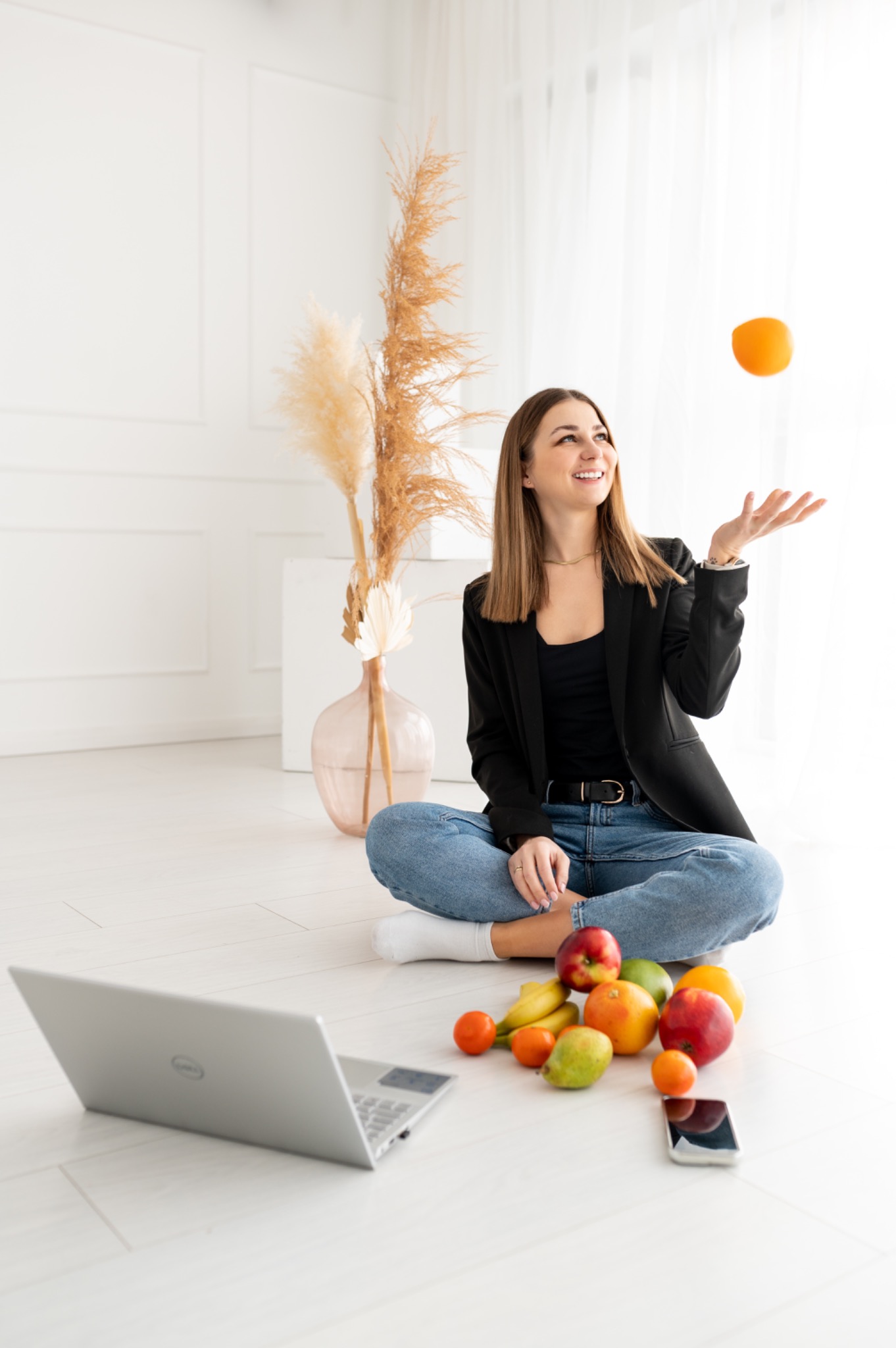 Uśmiechnięta kobieta w czarnej marynarce żongluje pomarańczą siedząc na podłodze z laptopem i owocami w jasnym, minimalistycznym wnętrzu.