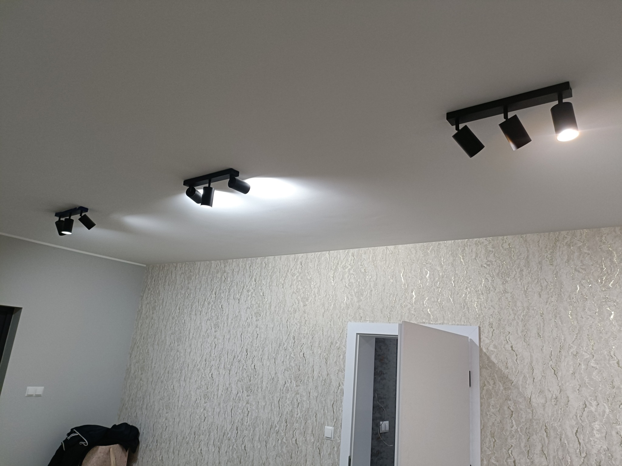 Nowoczesne oświetlenie sufitowe z trzema czarnymi reflektorami punktowymi, skierowanymi w różnych kierunkach, na tle wzorzystej tapety i częściowo otwartych białych drzwi.