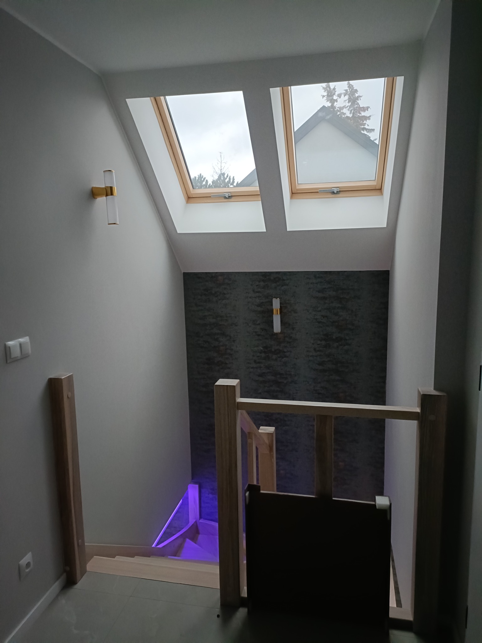 Nowoczesne schody z drewnianą balustradą i oświetleniem LED w kolorze fioletowym, widoczne dwa okna dachowe w skosie, ściana z ciemną tapetą o wzorze imitującym kamień, minimalistyczne kinkiety po...