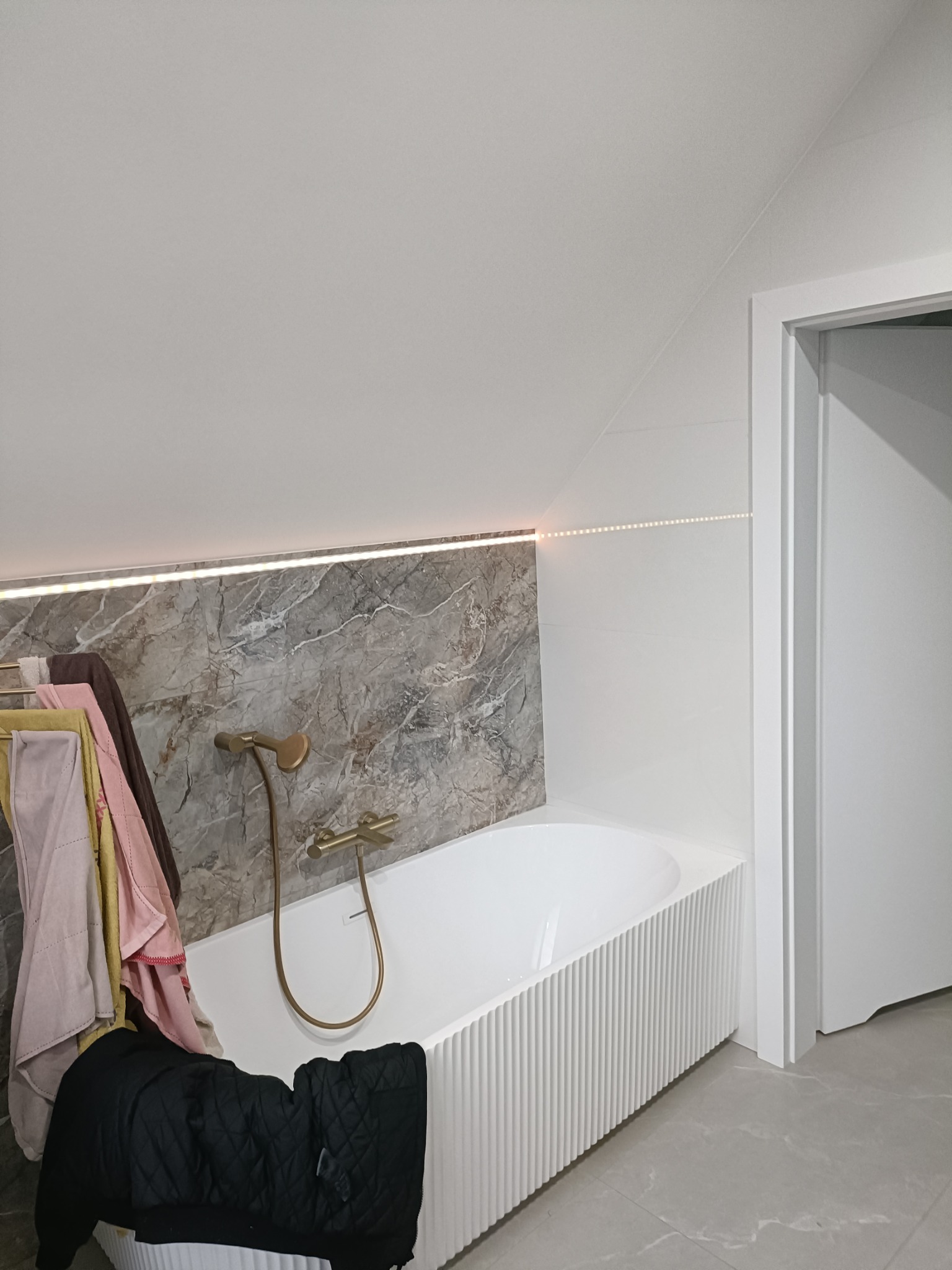 Nowoczesna łazienka na poddaszu z wanną obudowaną białymi lamelami, bateria w kolorze szczotkowanego złota, marmurowe płytki podświetlone listwą LED, ręczniki na wieszaku i czarna kurtka.