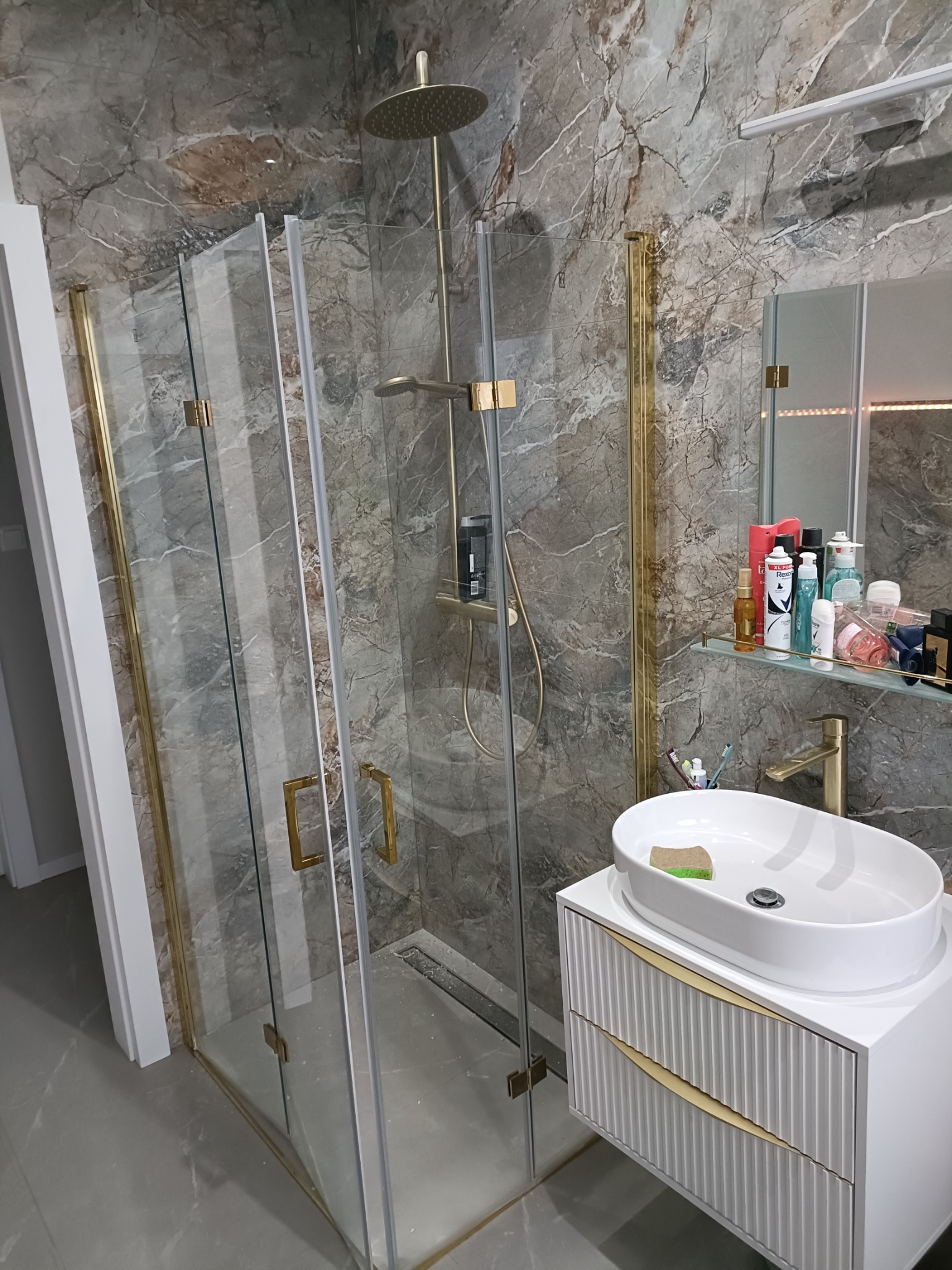 Nowoczesna łazienka z kabiną prysznicową ze złotymi ramami, umywalką nablatową na białej szafce z frezowanymi frontami i marmurowymi płytkami na ścianach.