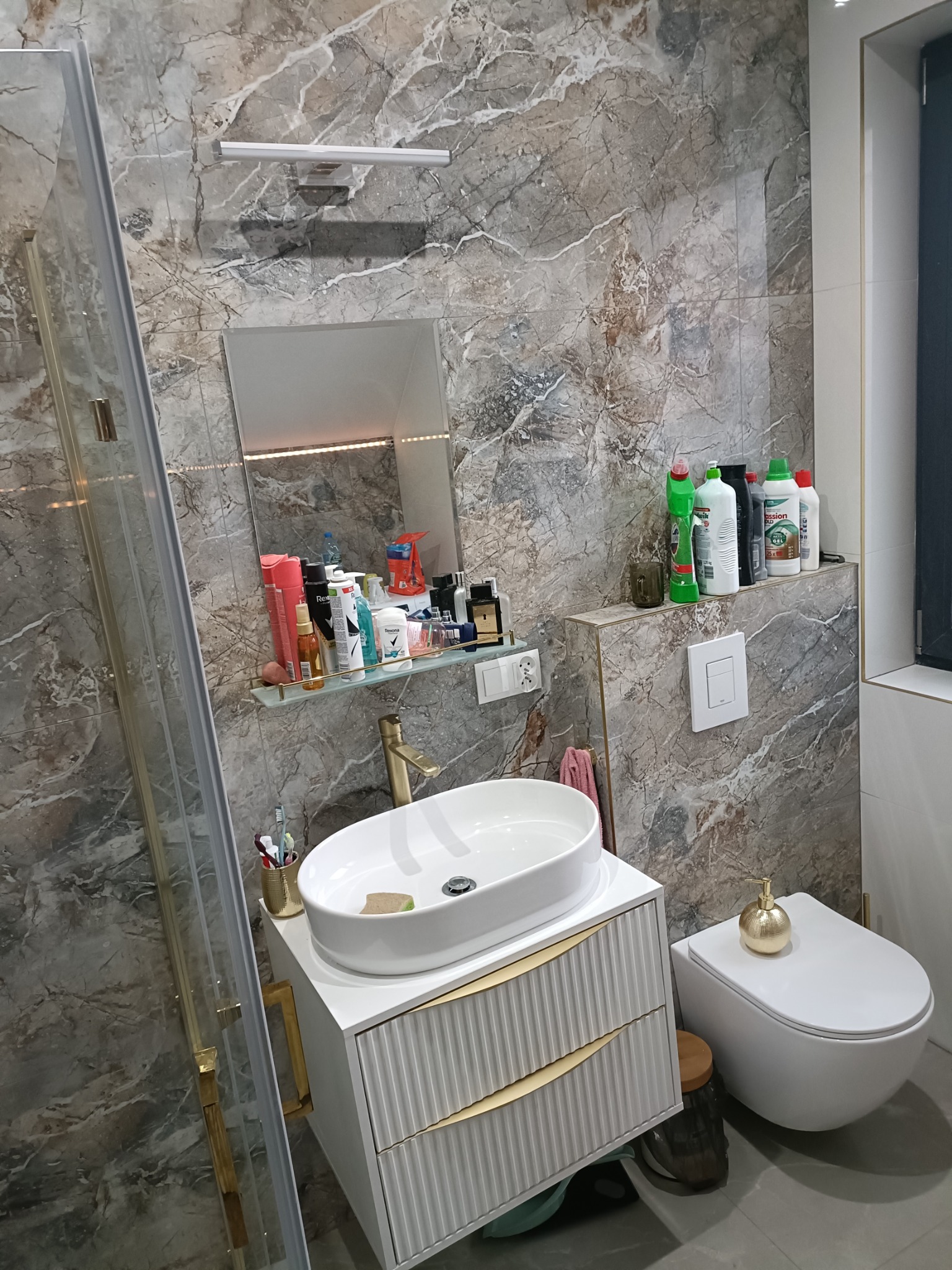 Nowoczesna łazienka po remoncie: marmurowe ściany, biała umywalka nablatowa z szafką w kolorze złamanej bieli, złota bateria, toaleta z dekoracyjną złotą buteleczką na zbiorniku, szklana kabina...
