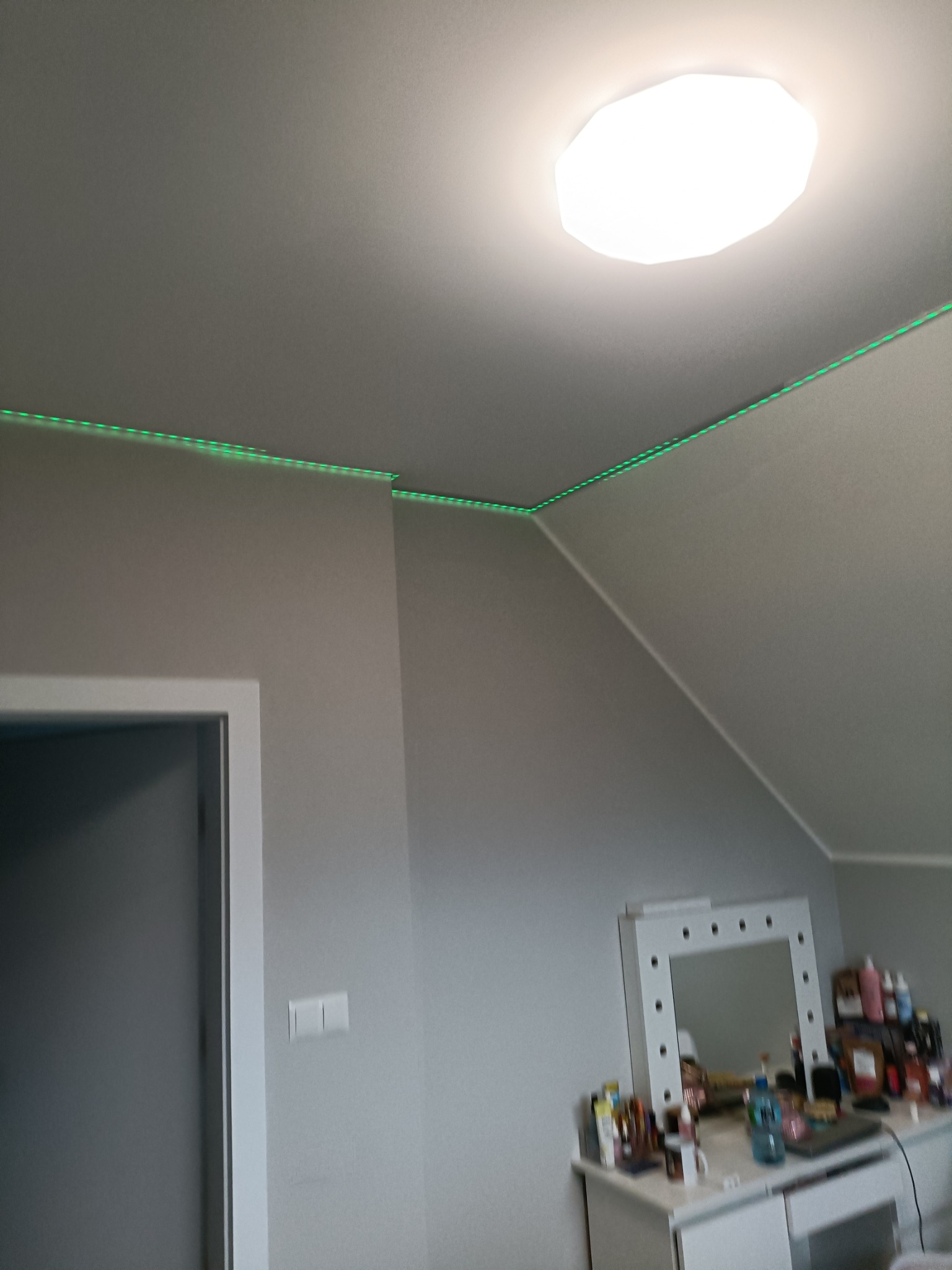 Szare wnętrze pokoju na poddaszu z oświetleniem LED wzdłuż krawędzi sufitu, widoczna lampa sufitowa i toaletka z lustrem.