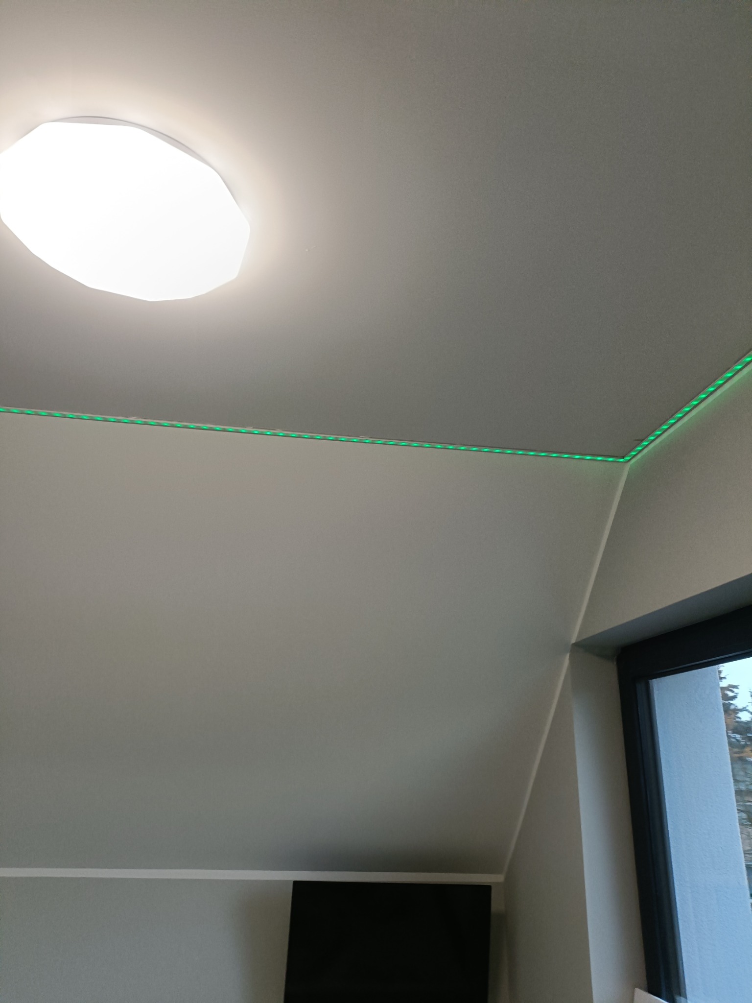Skośny sufit poddasza z zamontowanym oświetleniem LED wzdłuż krawędzi, widoczna okrągła lampa sufitowa i fragment okna.