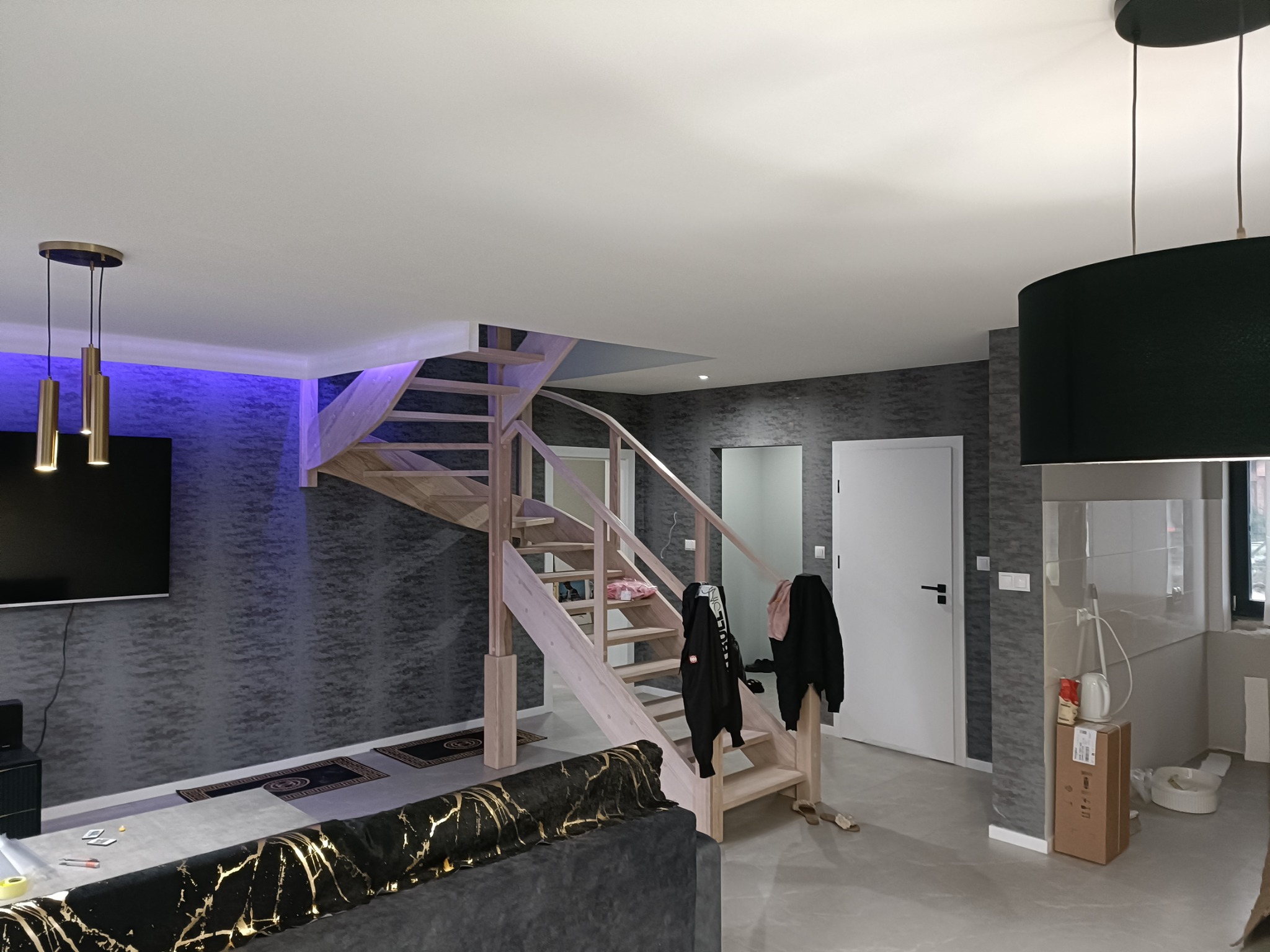 Wnętrze domu z drewnianymi schodami, szarą tapetą i elementami wykończenia w trakcie realizacji, widoczne ubrania na schodach i lampy sufitowe.