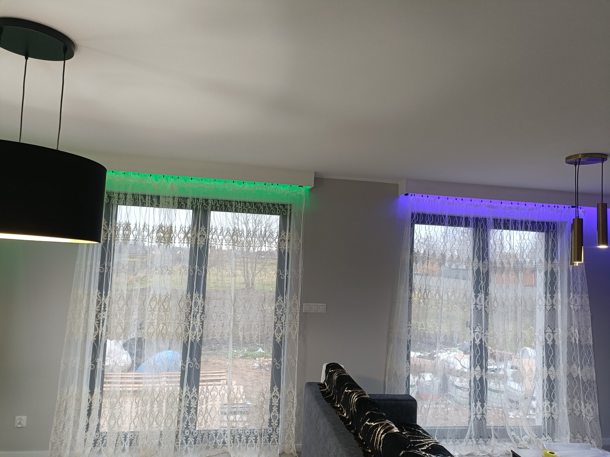 Salon z oknami udekorowanymi białymi firanami, podświetlonymi od góry zielonym i fioletowym światłem LED, widoczne lampy wiszące i fragment szarej sofy.