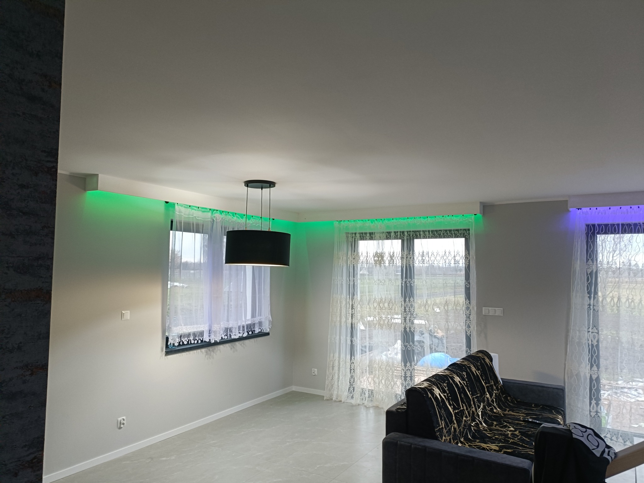 Nowoczesny salon z szarą sofą z wzorem marmuru, oświetleniem LED w kolorze zielonym i fioletowym nad oknami z białymi zasłonami oraz wiszącą lampą z czarnym abażurem.