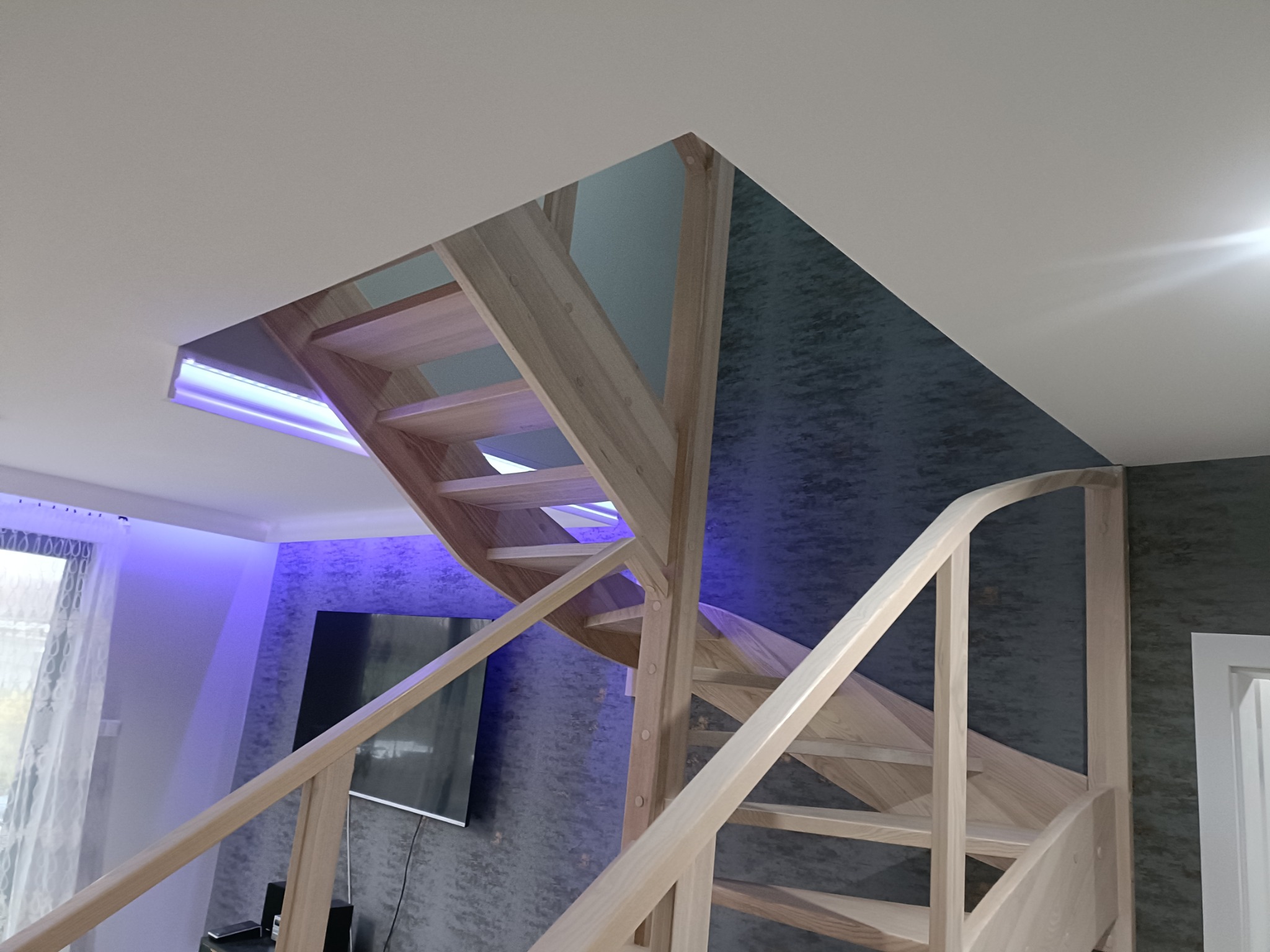 Jasne drewniane schody z oświetleniem LED wkomponowanym w sufit, widok z dołu na konstrukcję i dekoracyjną ścianę w tle.