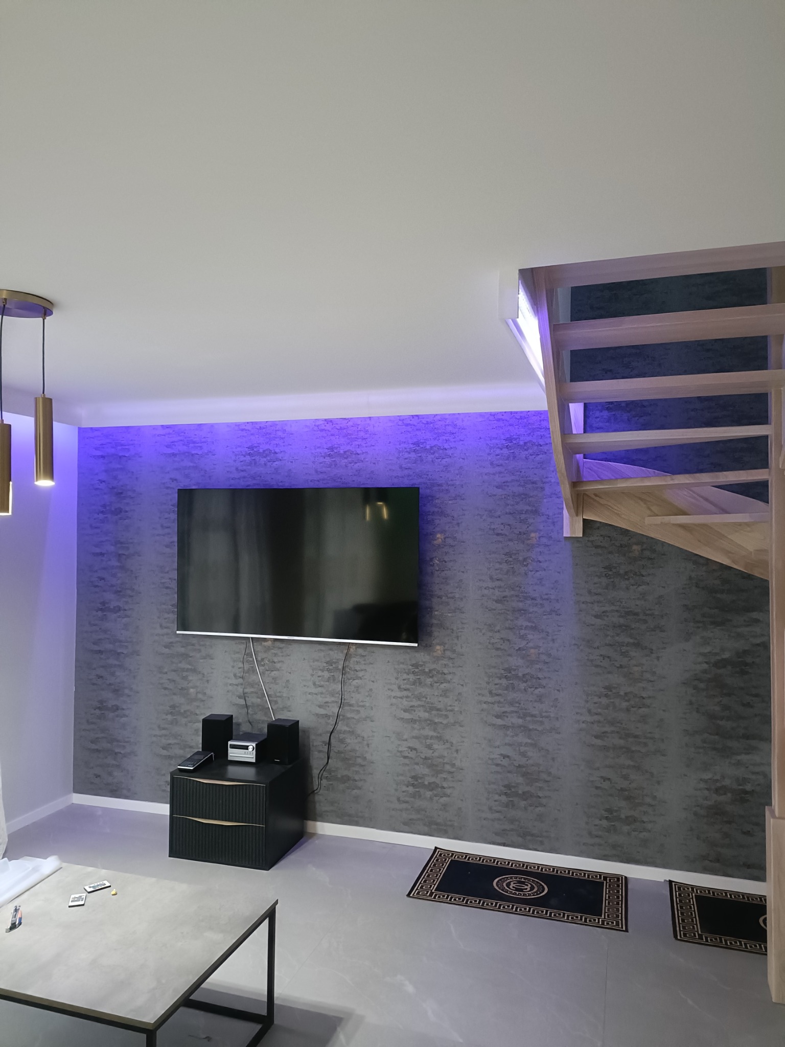 Nowoczesny salon z podświetloną ścianą, telewizorem, schodami drewnianymi i ciemnymi dywanikami z greckim wzorem.