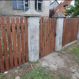 Mystol Pawel kowalski - Drewniane ogrodzenie z furtką, brązowe deski pionowe, szare betonowe słupki z daszkami, tło: budynek z szarej cegły i dom z czerwonym dachem.