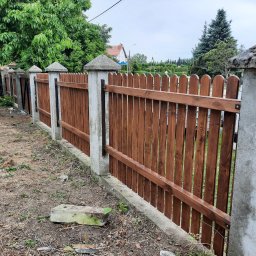 Mystol Pawel kowalski - Drewniane ogrodzenie z betonowymi słupkami i daszkami, pomalowane na brązowo, z widocznymi elementami montażowymi, otoczone zielenią. Widok z perspektywy.