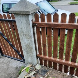 Mystol Pawel kowalski - Świeżo pomalowane drewniane ogrodzenie z bramką, widoczne słupki betonowe i fragment samochodu w tle. Ujęcie z niskiej perspektywy.