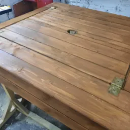 Drewniany stół z otworem na parasol, wykonany z desek w ciepłym, brązowym odcieniu, wsparty na drewnianej konstrukcji. Widoczne zawiasy i ślady obróbki.