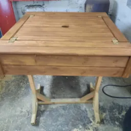 Stół drewniany z otwieranym blatem na zawiasach, prawdopodobnie do przechowywania. Widoczny otwór w blacie. Wykończenie bejcą w ciepłym odcieniu brązu. Proste nogi z drewna.