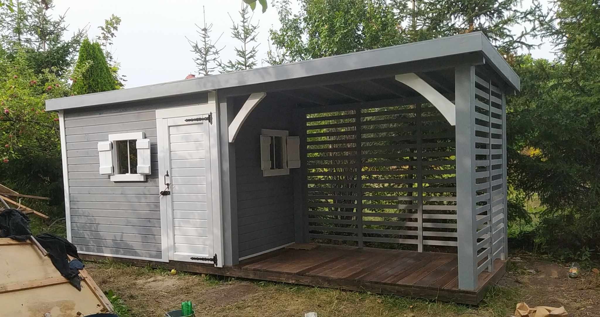 Szara konstrukcja z zadaszeniem, częściowo zabudowana, z drewnianą podłogą i ażurową ścianą, otoczona zielenią.