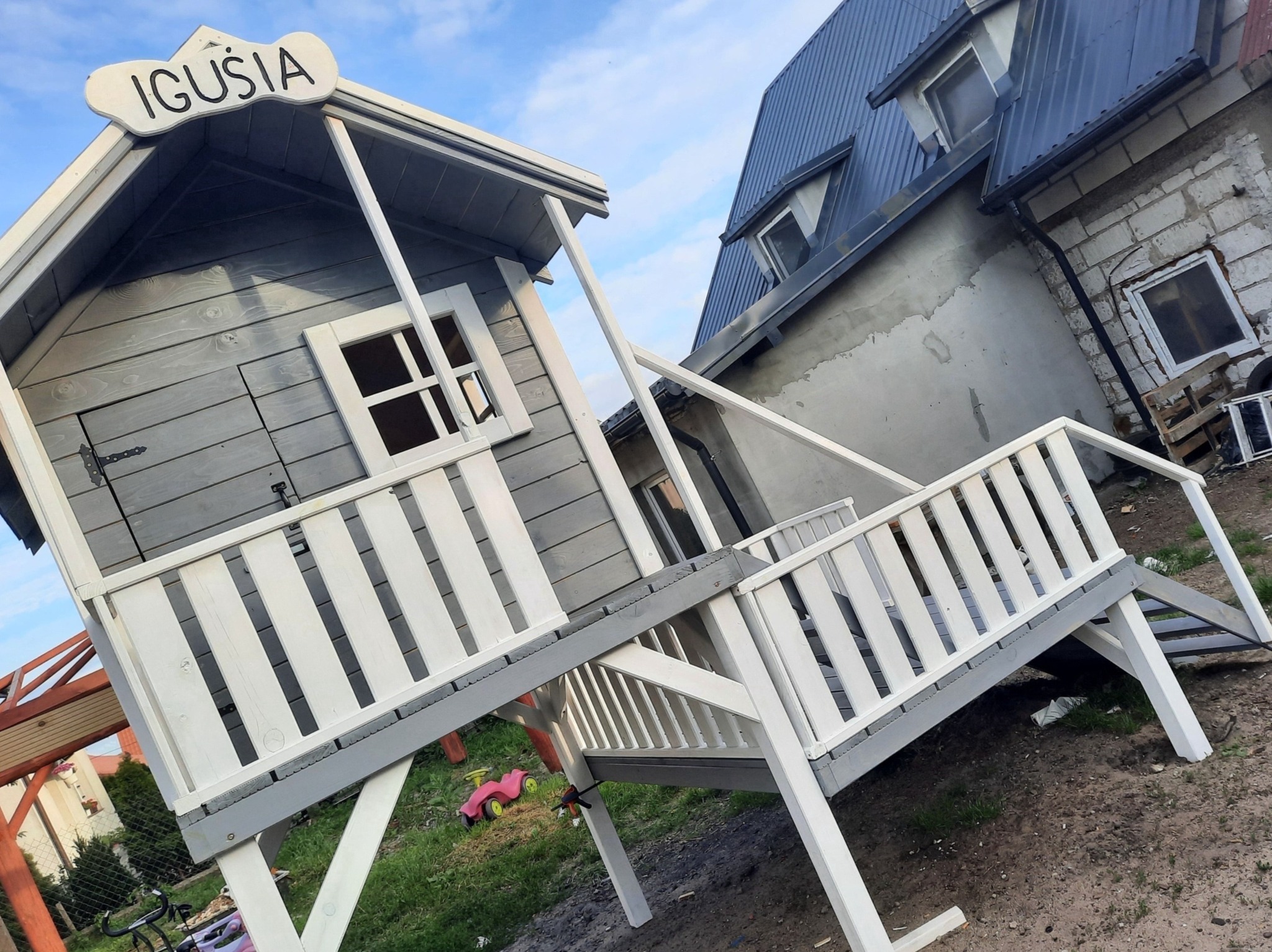 Szara drewniana konstrukcja placu zabaw z białymi balustradami i napisem 'Iguś' na tle niebieskiego nieba i budynku. Ujęcie z dołu, podkreślające wysokość konstrukcji.