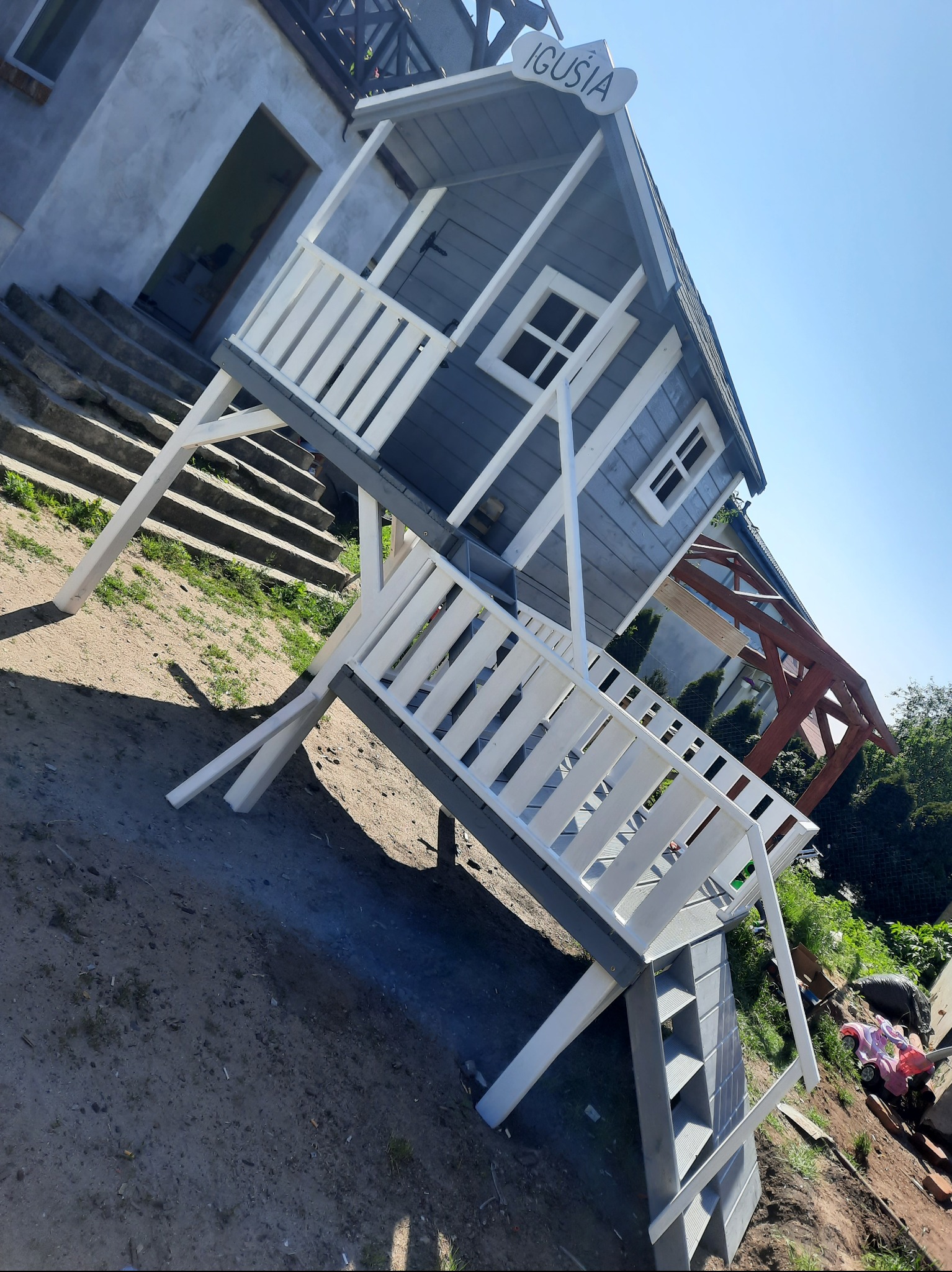 Nietypowy, szary domek dla dzieci z białymi balustradami i schodami, umieszczony na podwyższeniu, z tabliczką z imieniem 'Iguś' na dachu.