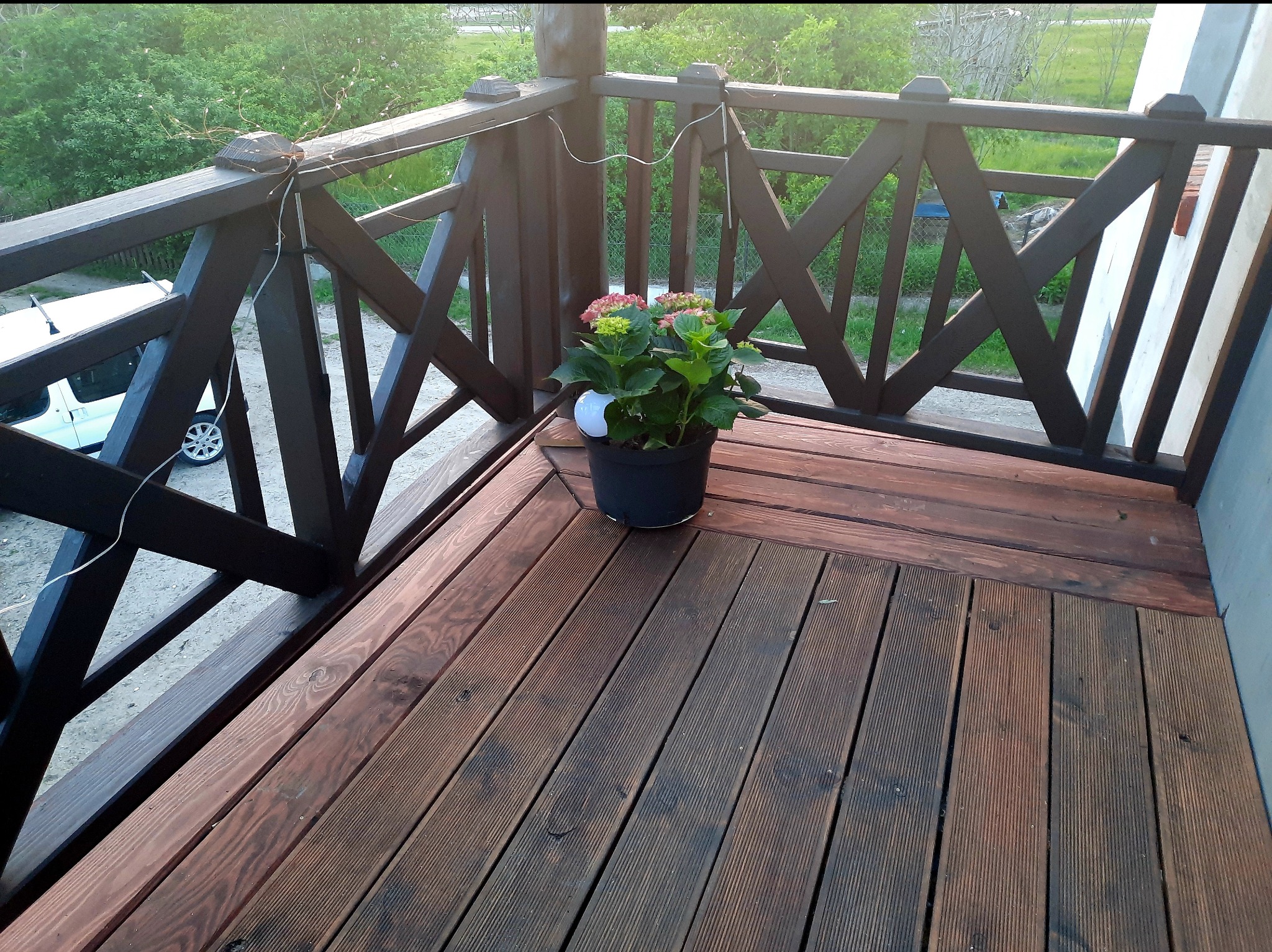 Kątowe ujęcie balkonu z drewnianą podłogą i balustradą w brązowym odcieniu. Na podłodze stoi doniczka z kwiatami. W tle zieleń.